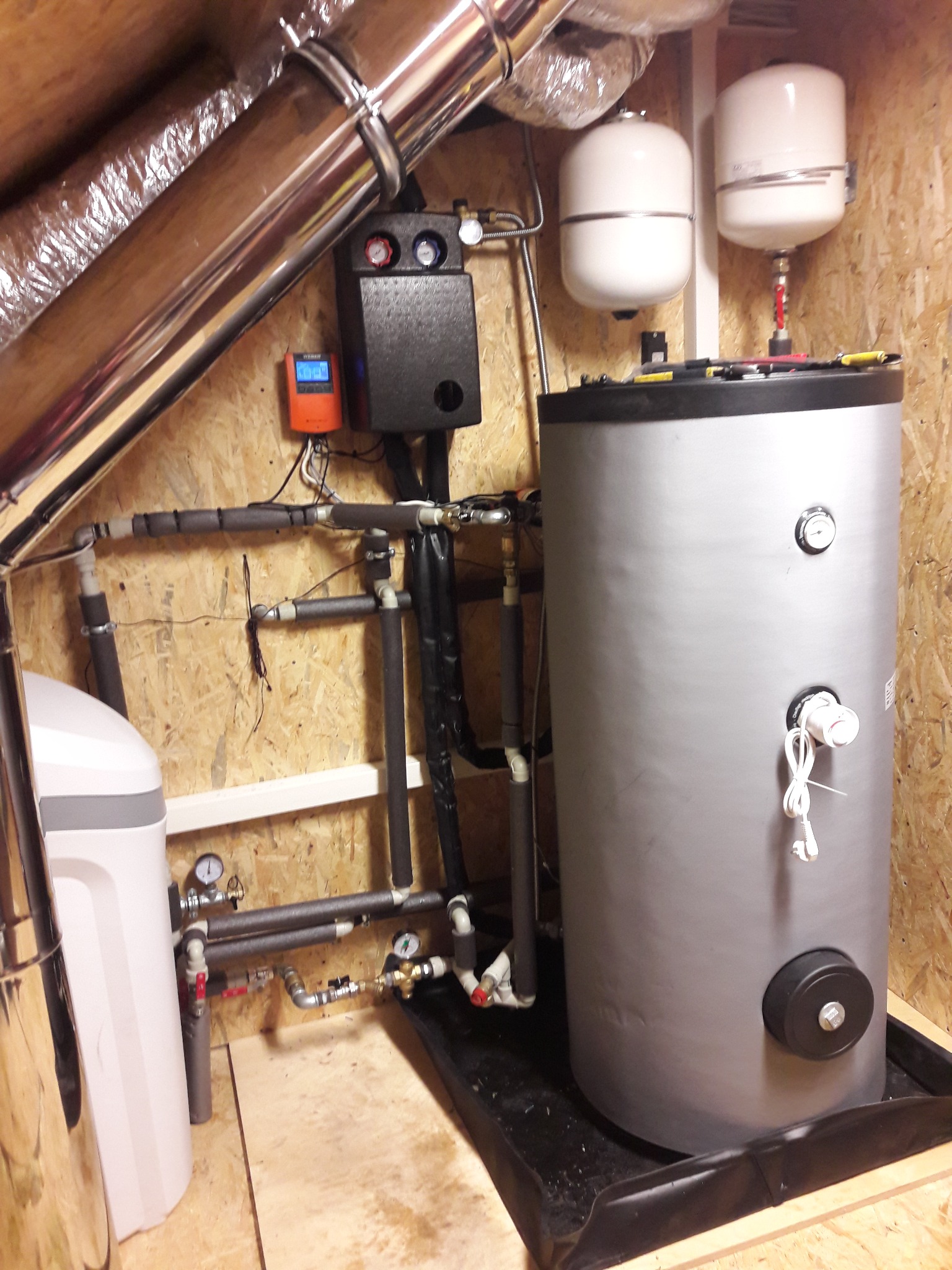 Szary bojler z podłączonymi rurami, manometrami i zabezpieczeniami, zainstalowany w pomieszczeniu ze ścianami wyłożonymi płytami OSB, widoczne elementy instalacji grzewczej i wodnej.