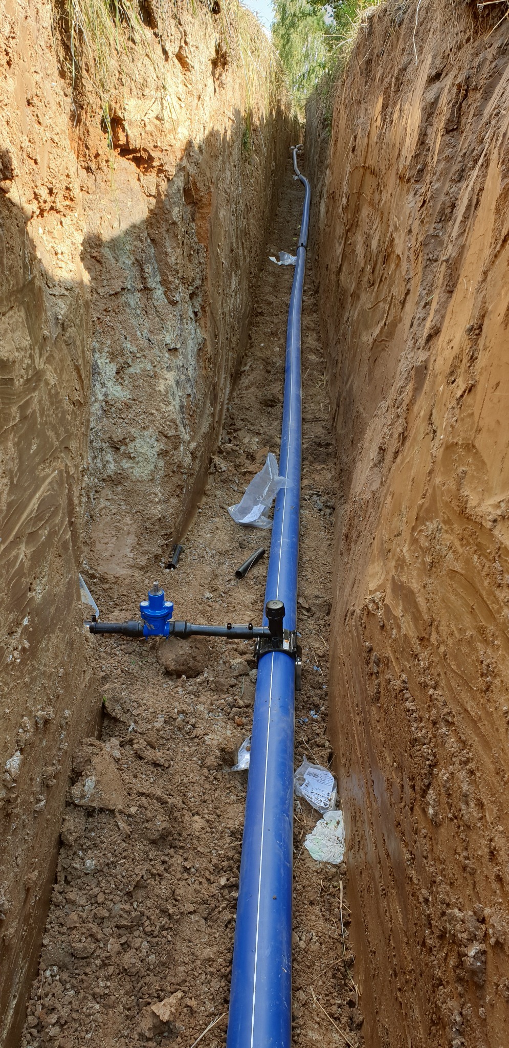 Niebieska rura wodociągowa z zaworem w wykopie ziemnym, widoczne elementy montażowe i resztki opakowań.