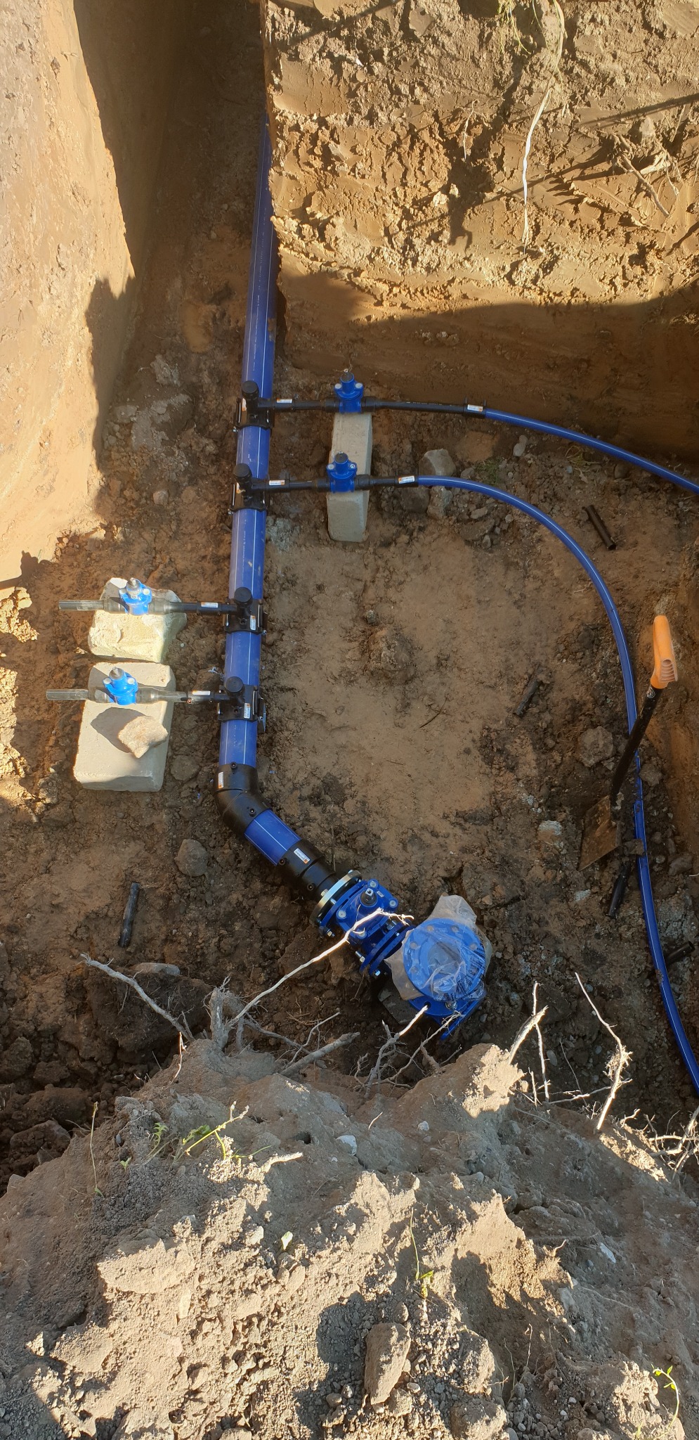 Niebieska instalacja wodna z zaworami i złączkami zamontowana w wykopie ziemnym, częściowo zabezpieczona folią, widoczna łopata w tle.