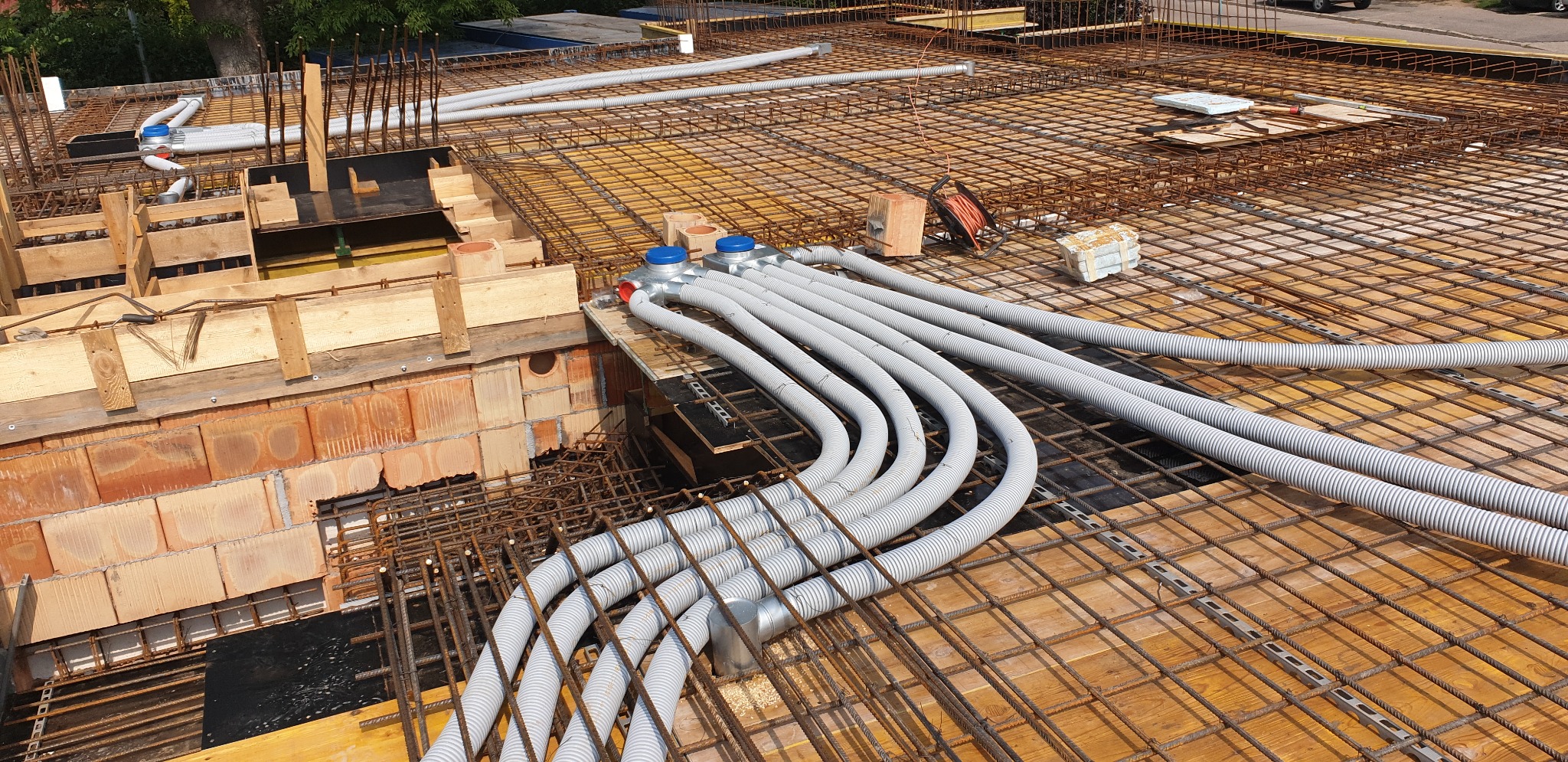Szare, karbowane rury ogrzewania podłogowego z niebieskimi rozdzielaczami ułożone na siatce zbrojeniowej w surowym stanie deweloperskim budynku.