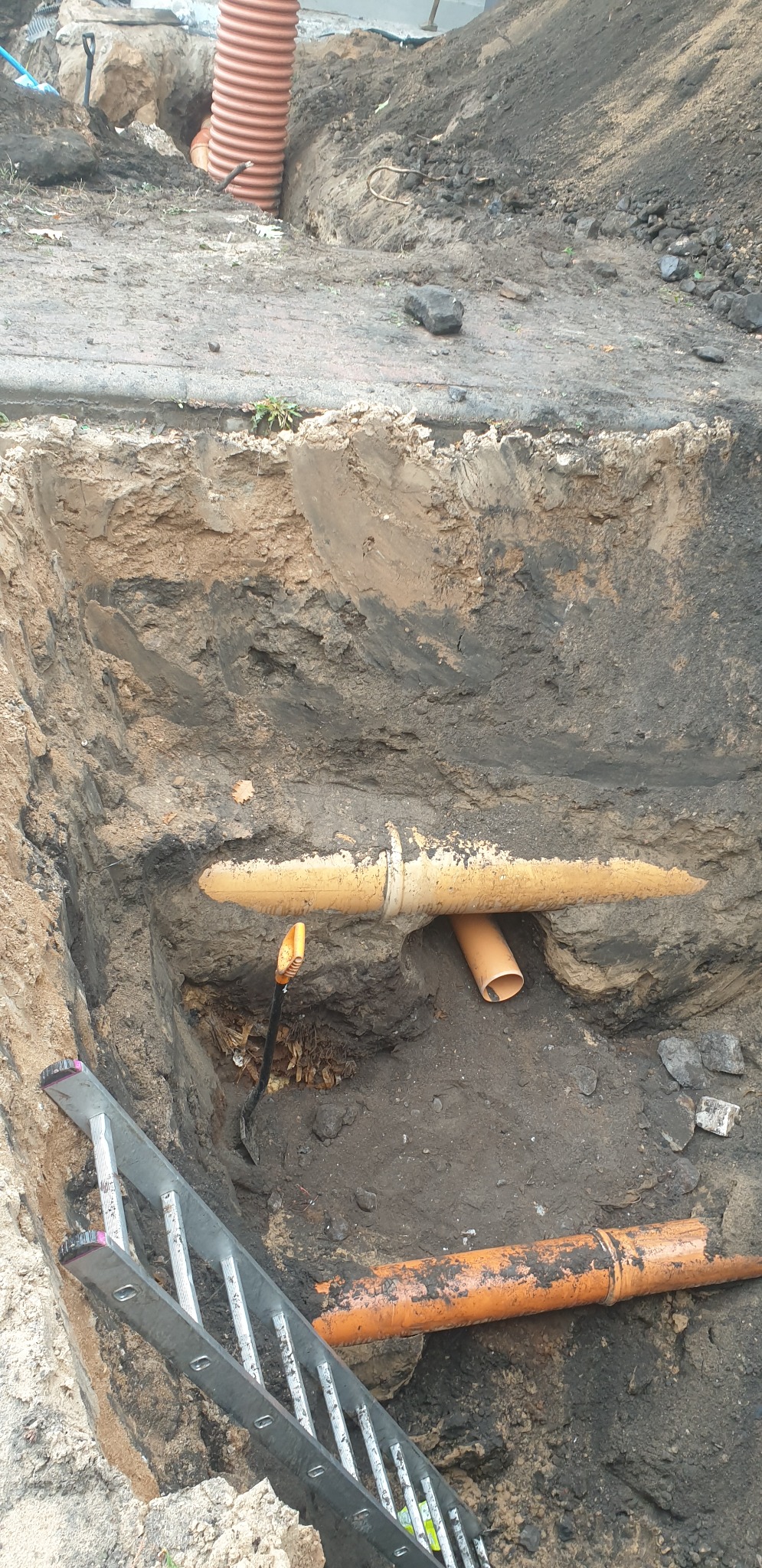 Wykop z widocznymi rurami kanalizacyjnymi z tworzywa sztucznego, łopatą w wykopie i fragmentem drabiny opartą o krawędź wykopu.