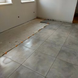 AB4 Artur Bukała - Układanie szarych płytek podłogowych w trakcie remontu w Piławie Dolnej, widoczne pomarańczowe klipsy poziomujące i fragment surowej wylewki.