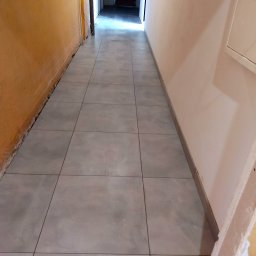 AB4 Artur Bukała - Korytarz w trakcie prac wykończeniowych, szare płytki podłogowe ułożone, ściany częściowo przygotowane do malowania, widoczne przewody i elementy instalacji elektrycznej.