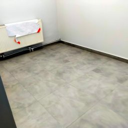 AB4 Artur Bukała - Puste, odświeżone pomieszczenie z szarymi płytkami podłogowymi, białe ściany z listwami przypodłogowymi, grzejnik z przyczepionym planem/rysunkiem z czerwonymi elementami.