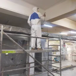 Pracownik w kombinezonie ochronnym, stojący na rusztowaniu, maluje sufit za pomocą pistoletu natryskowego w pomieszczeniu zabezpieczonym folią.