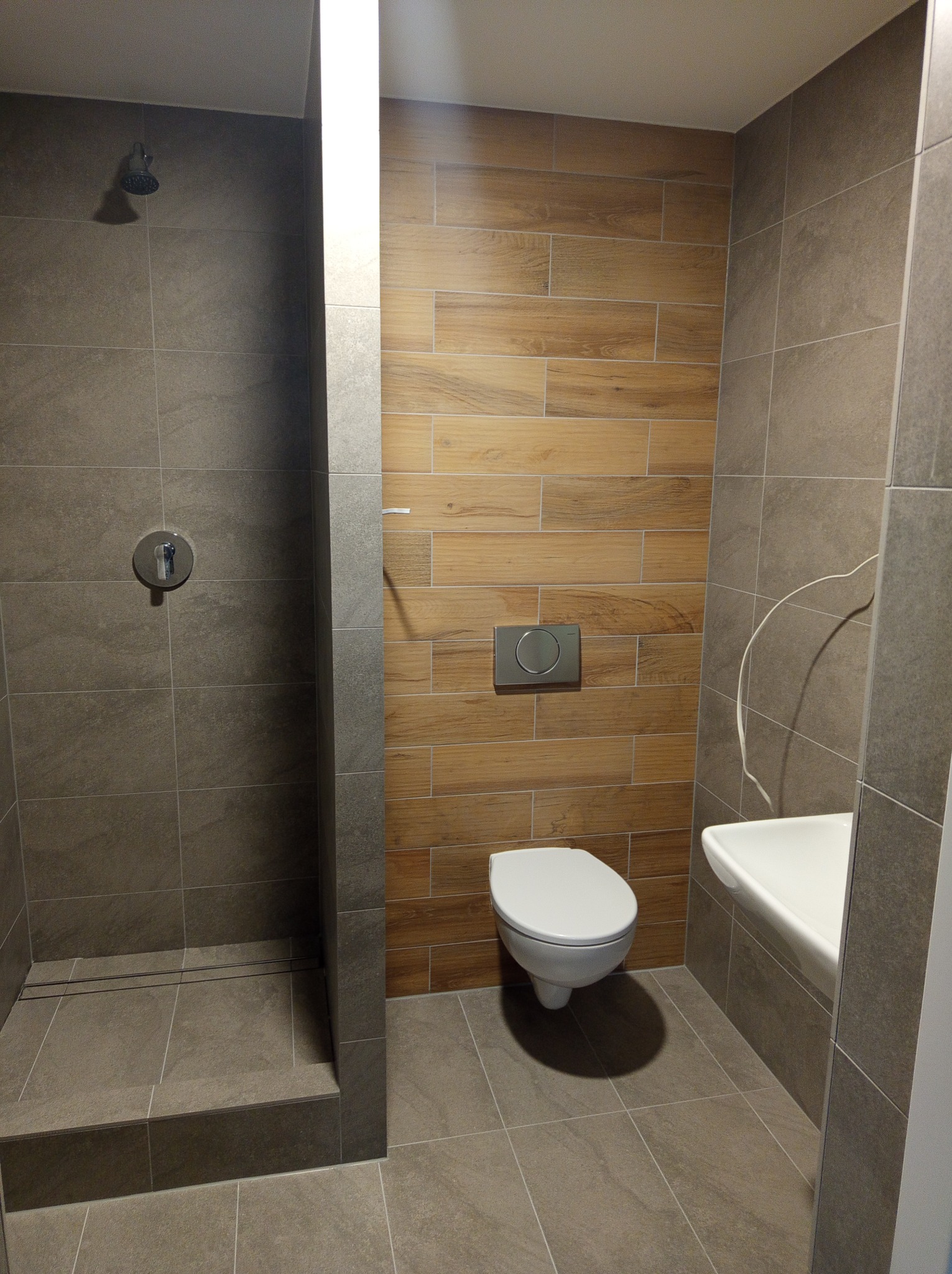 Nowoczesna łazienka z kabiną prysznicową typu walk-in, wiszącą toaletą i umywalką w rogu, wykończona płytkami w odcieniach szarości i drewna, z widoczną armaturą.