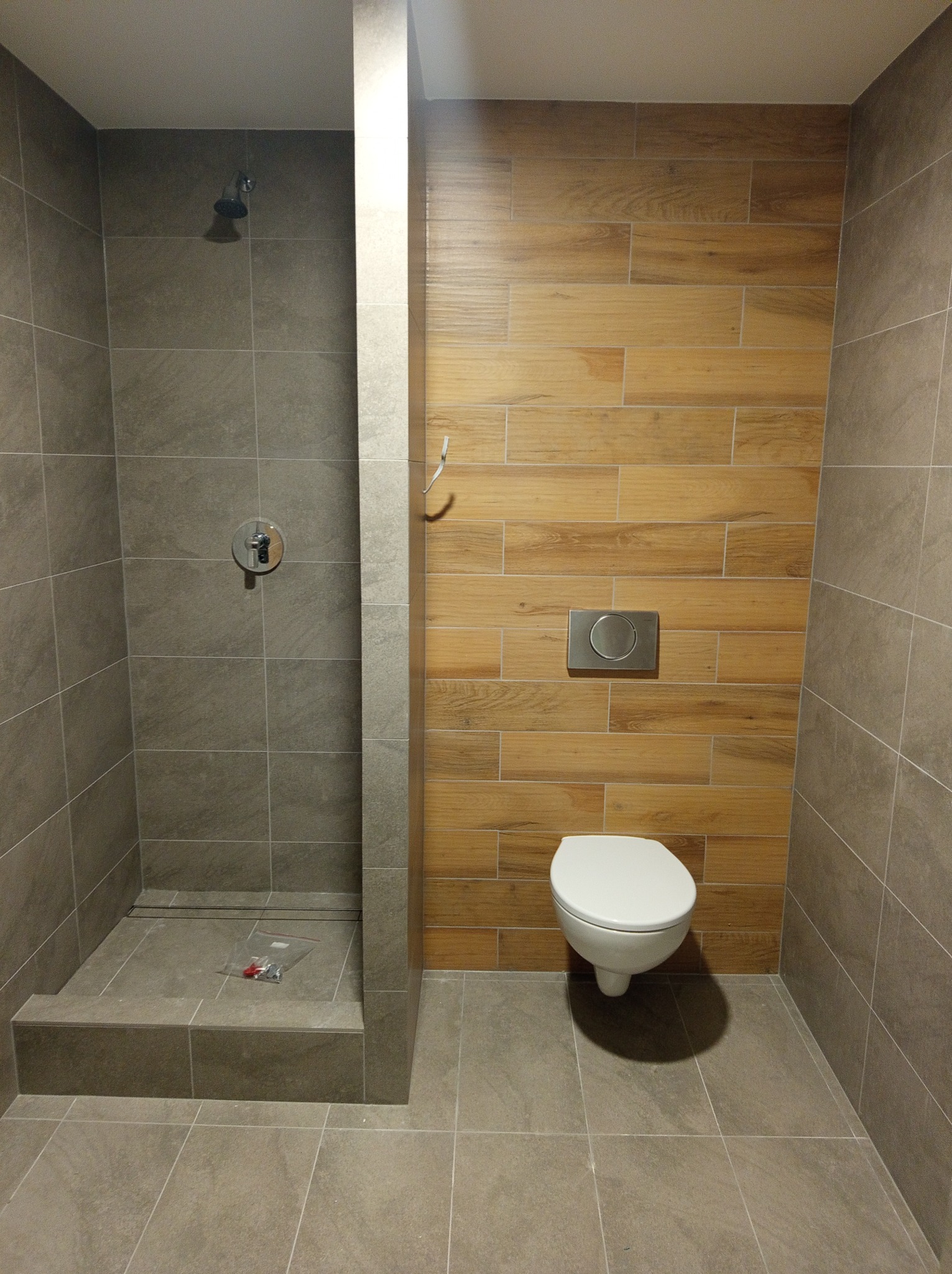 Nowoczesna łazienka z kabiną prysznicową wyłożoną szarą płytką i toaletą podwieszaną na ścianie z drewnopodobnych paneli; w kabinie prysznicowej leżą akcesoria montażowe w plastikowej torebce.