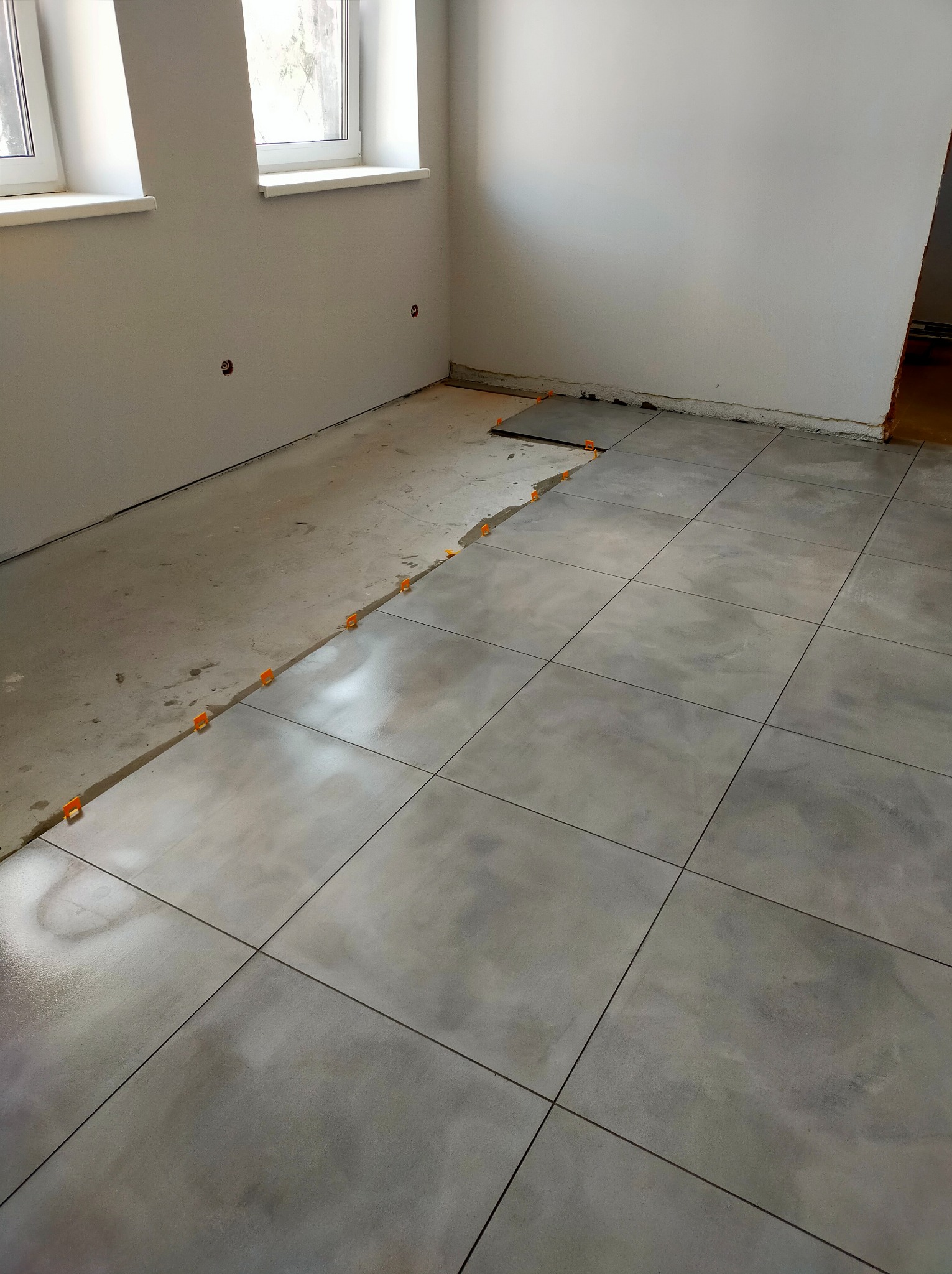 Układanie szarych płytek podłogowych w trakcie remontu w Piławie Dolnej, widoczne pomarańczowe klipsy poziomujące i fragment surowej wylewki.