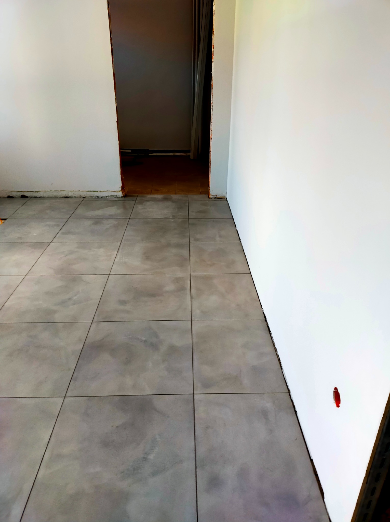 Szare płytki podłogowe ułożone w nowym pomieszczeniu z białymi ścianami, widoczny fragment czerwonego znacznika na ścianie.