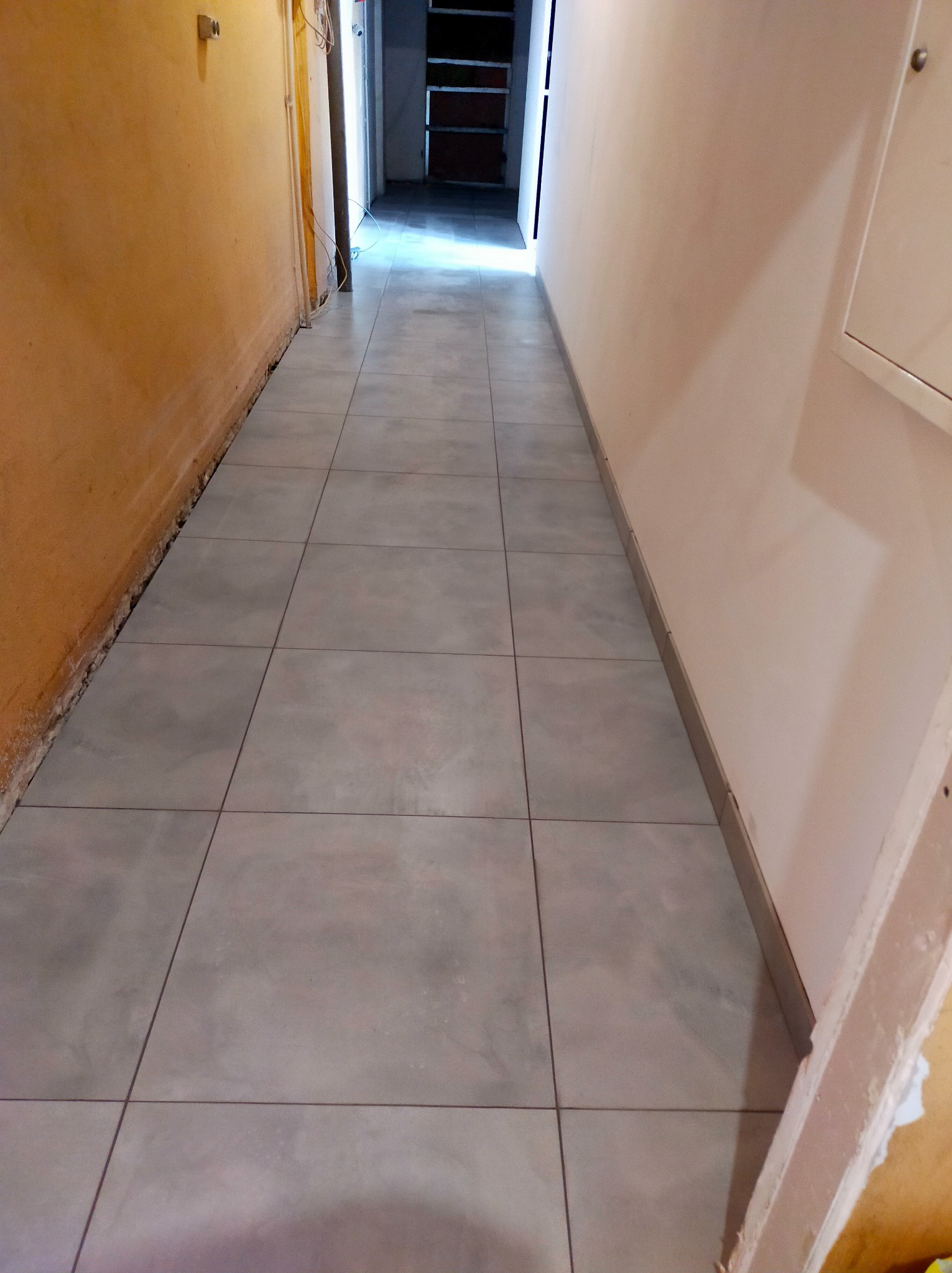 Korytarz w trakcie prac wykończeniowych, szare płytki podłogowe ułożone, ściany częściowo przygotowane do malowania, widoczne przewody i elementy instalacji elektrycznej.