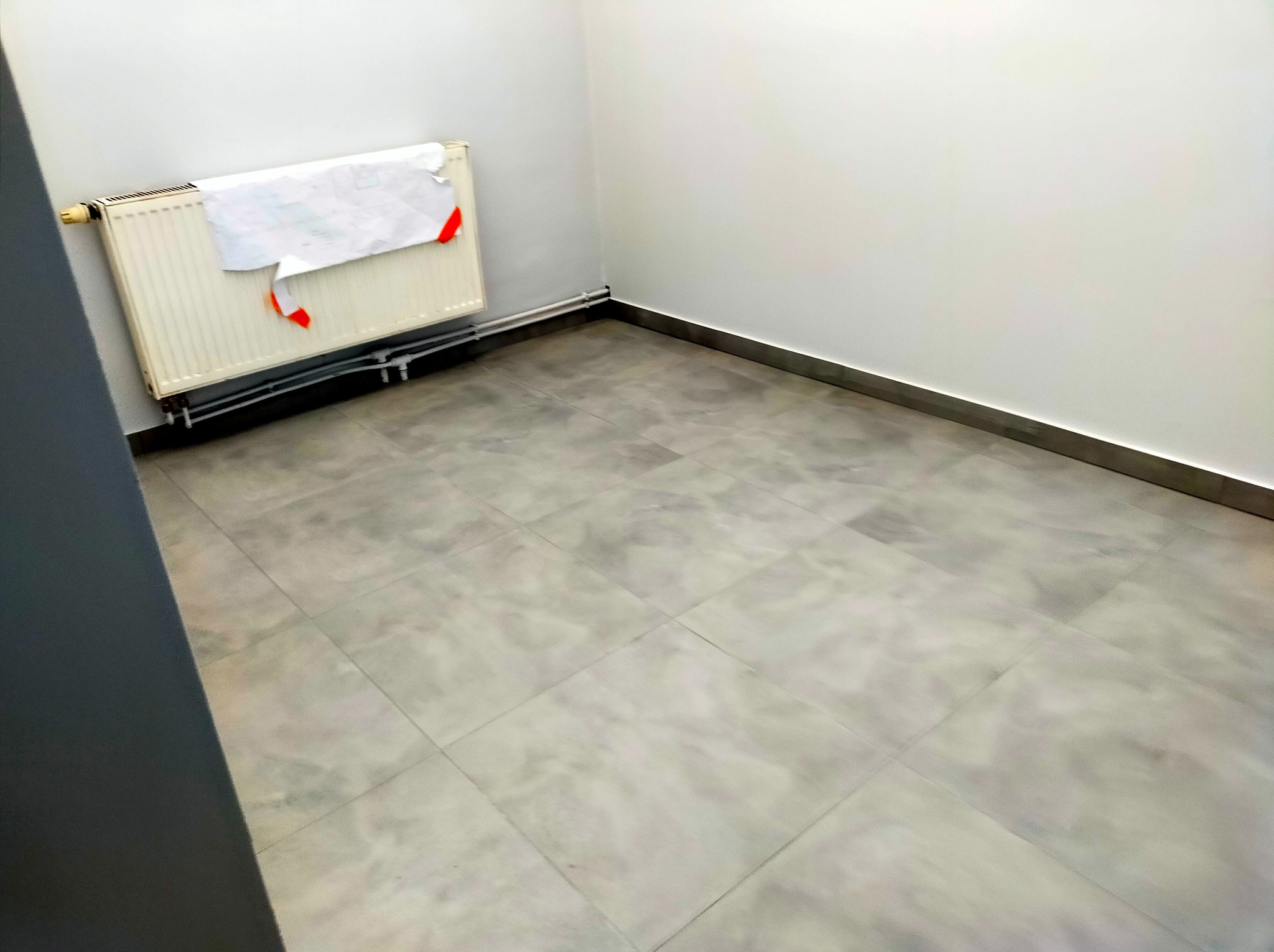 Puste, odświeżone pomieszczenie z szarymi płytkami podłogowymi, białe ściany z listwami przypodłogowymi, grzejnik z przyczepionym planem/rysunkiem z czerwonymi elementami.