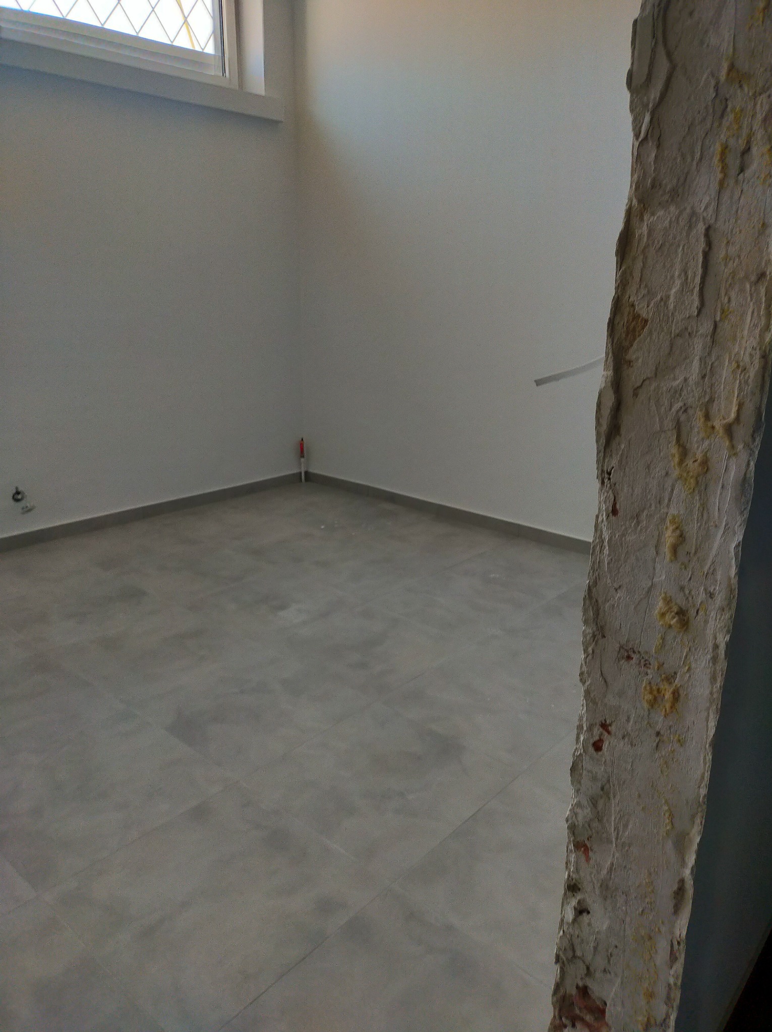 Wnętrze w trakcie prac remontowych: szara podłoga z płytek, fragment ściany z odsłoniętą cegłą i pianką montażową, gładkie, pomalowane na biało ściany w tle, mała rurka wystająca ze ściany.