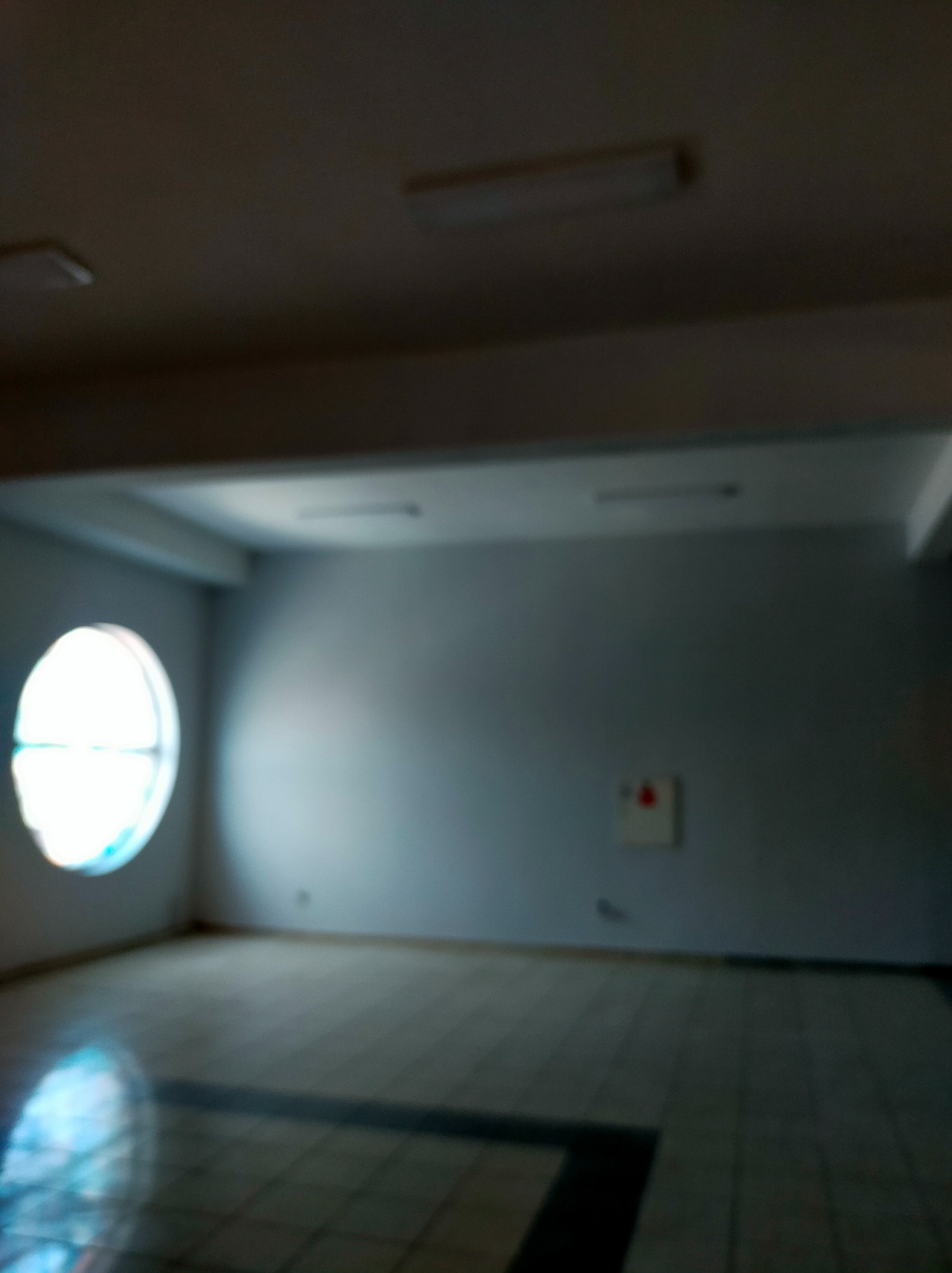 Puste, jasne pomieszczenie biurowe z okrągłym oknem, białe ściany i płytki na podłodze, widoczny panel przeciwpożarowy, rozproszone światło, nieostre zdjęcie.