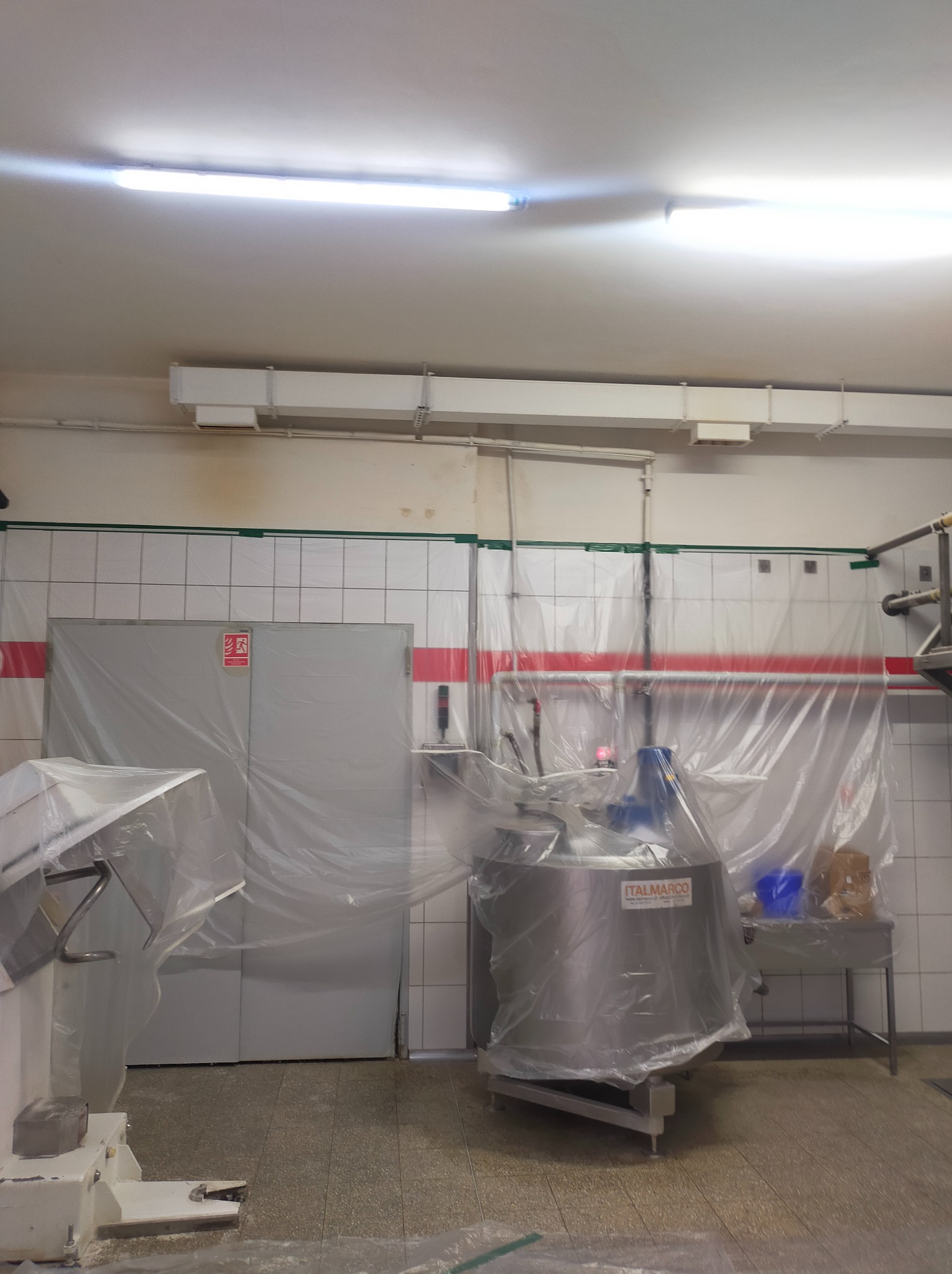 Przygotowanie do prac w pomieszczeniu przemysłowym: zabezpieczone folią ściany z białych płytek i stalowy zbiornik Italmarco z niebieską pokrywą, podłużna lampa na suficie.