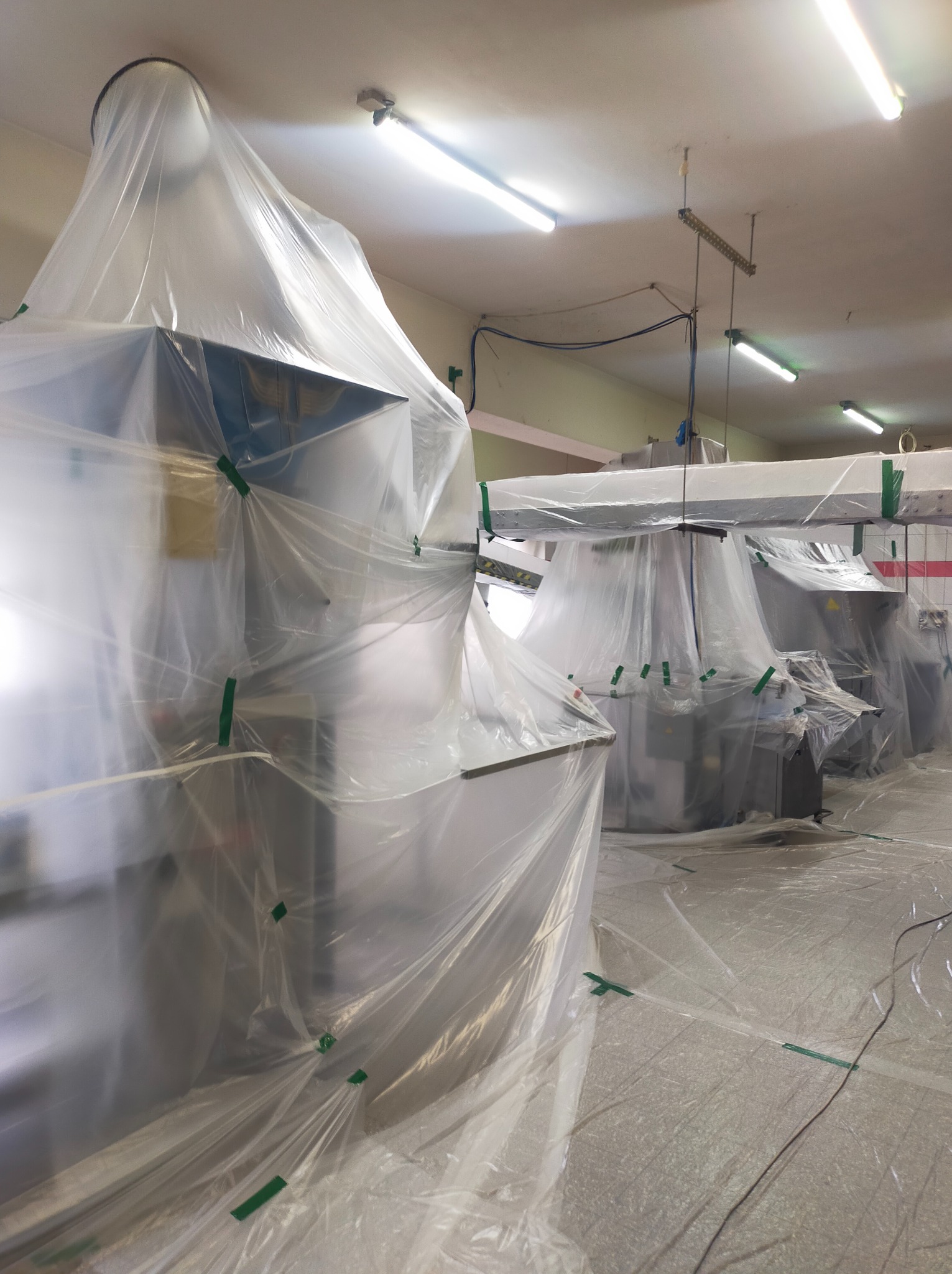 Przygotowanie do malowania w pomieszczeniu biurowym: maszyny i podłoga zabezpieczone folią ochronną, przymocowaną zieloną taśmą malarską.