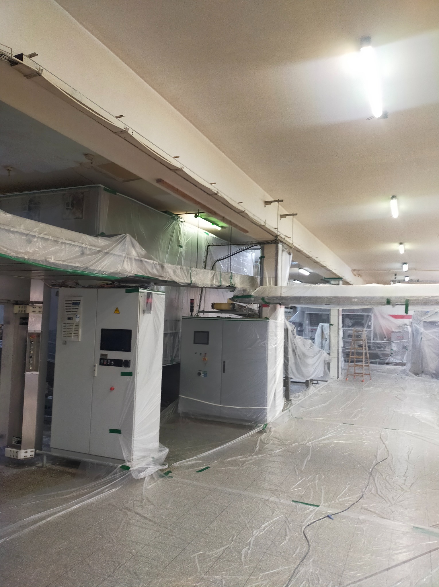 Przygotowanie do prac malarskich w hali produkcyjnej: maszyny i podłoga zabezpieczone folią ochronną, widoczne szafy sterownicze i drabina.