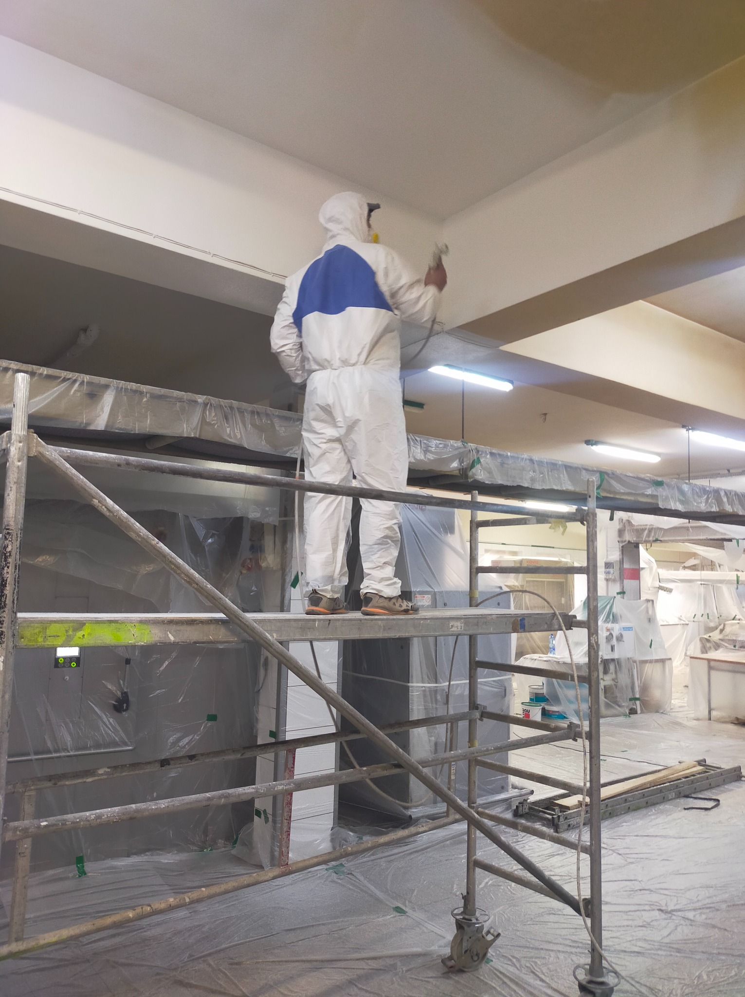 Pracownik w kombinezonie ochronnym, stojący na rusztowaniu, maluje sufit za pomocą pistoletu natryskowego w pomieszczeniu zabezpieczonym folią.