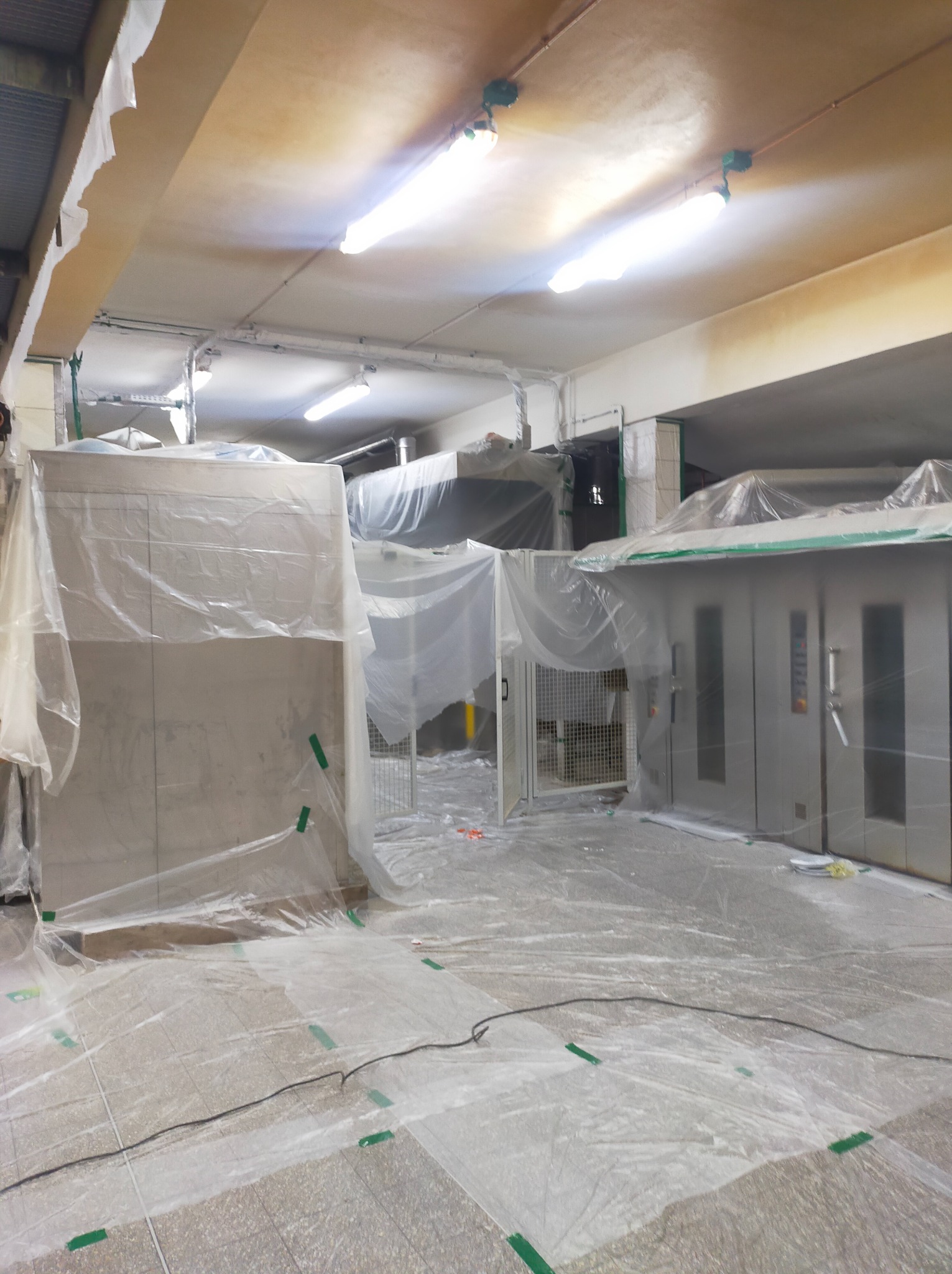 Przygotowanie do prac w pomieszczeniu przemysłowym, zabezpieczone folią malarską ściany, podłoga i urządzenia, widoczne zielone taśmy mocujące.
