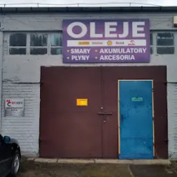 Fasada warsztatu z dużym szyldem reklamującym oleje, smary, akumulatory, płyny i akcesoria; brązowa brama garażowa i niebieskie drzwi wejściowe.