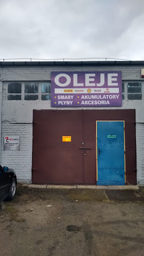 Fasada warsztatu z dużym szyldem reklamującym oleje, smary, akumulatory, płyny i akcesoria; brązowa brama garażowa i niebieskie drzwi wejściowe.