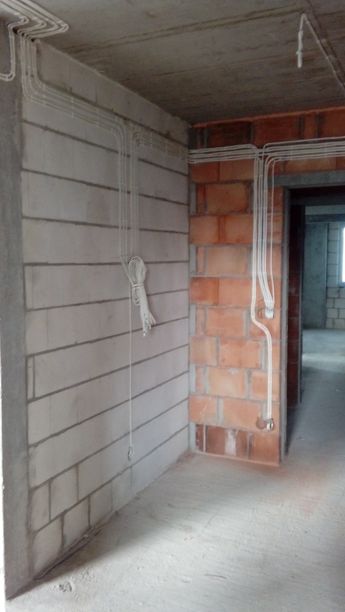 Surowe wnętrze w trakcie budowy z widocznymi przewodami elektrycznymi poprowadzonymi po ścianach z betonu komórkowego i cegły, zwisający pęk kabli, w tle widoczne kolejne pomieszczenia.