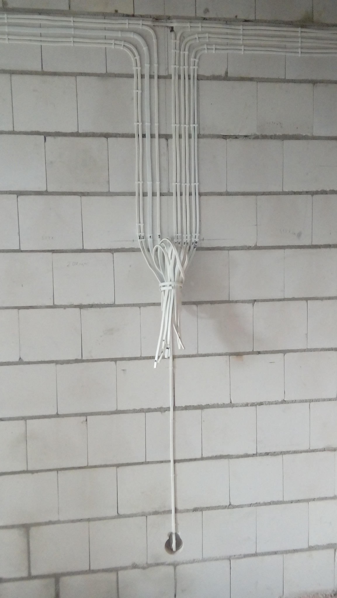 Białe kable elektryczne prowadzone po ścianie z białych bloczków betonowych, zebrane w wiązkę i spuszczone do otworu w ścianie.