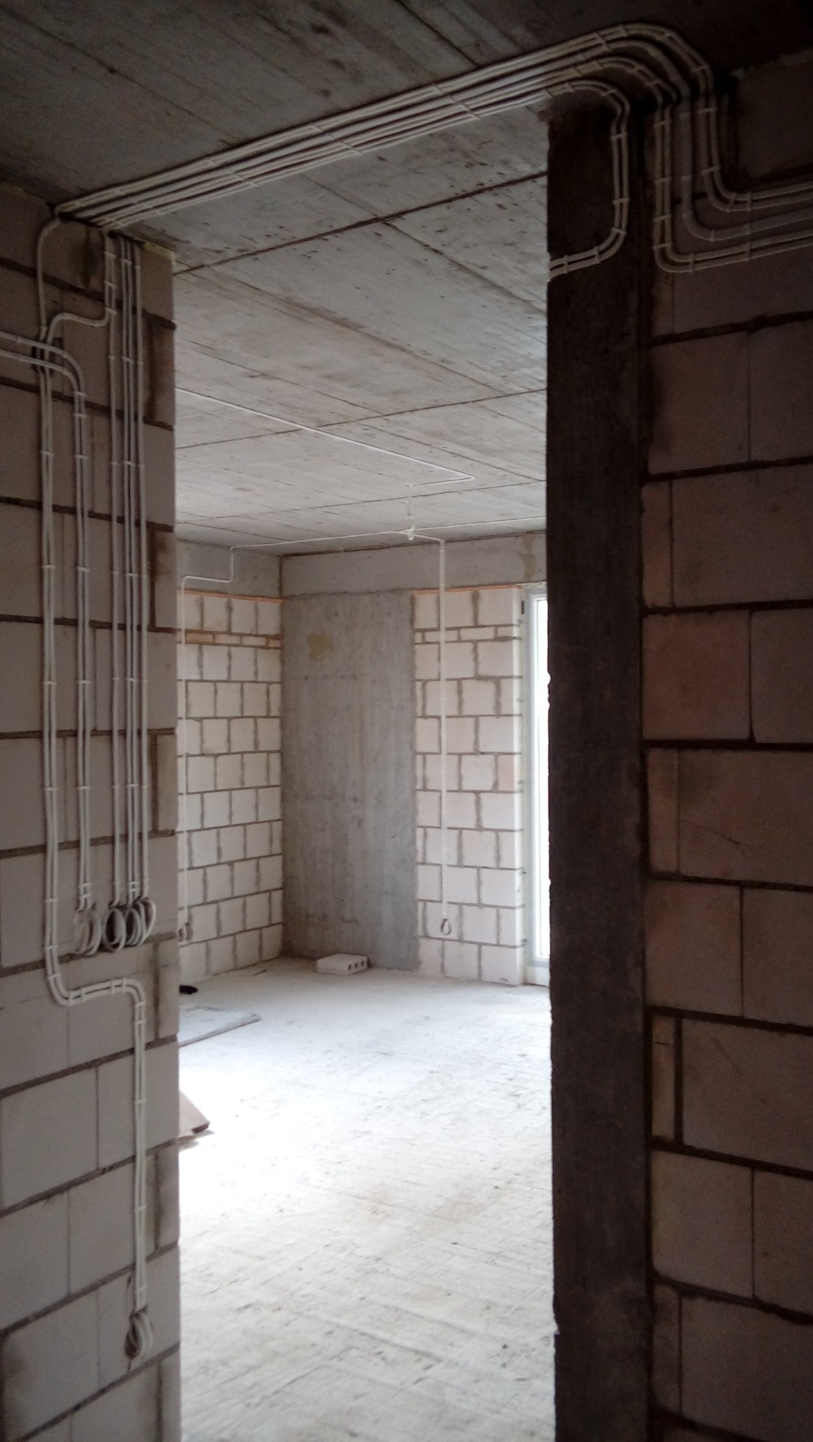 Surowe wnętrze w trakcie prac wykończeniowych, widoczne przewody elektryczne ułożone na ścianach i suficie, prowadzone w peszlach i listwach, ściany z białego bloczka, surowy betonowy strop, okno...