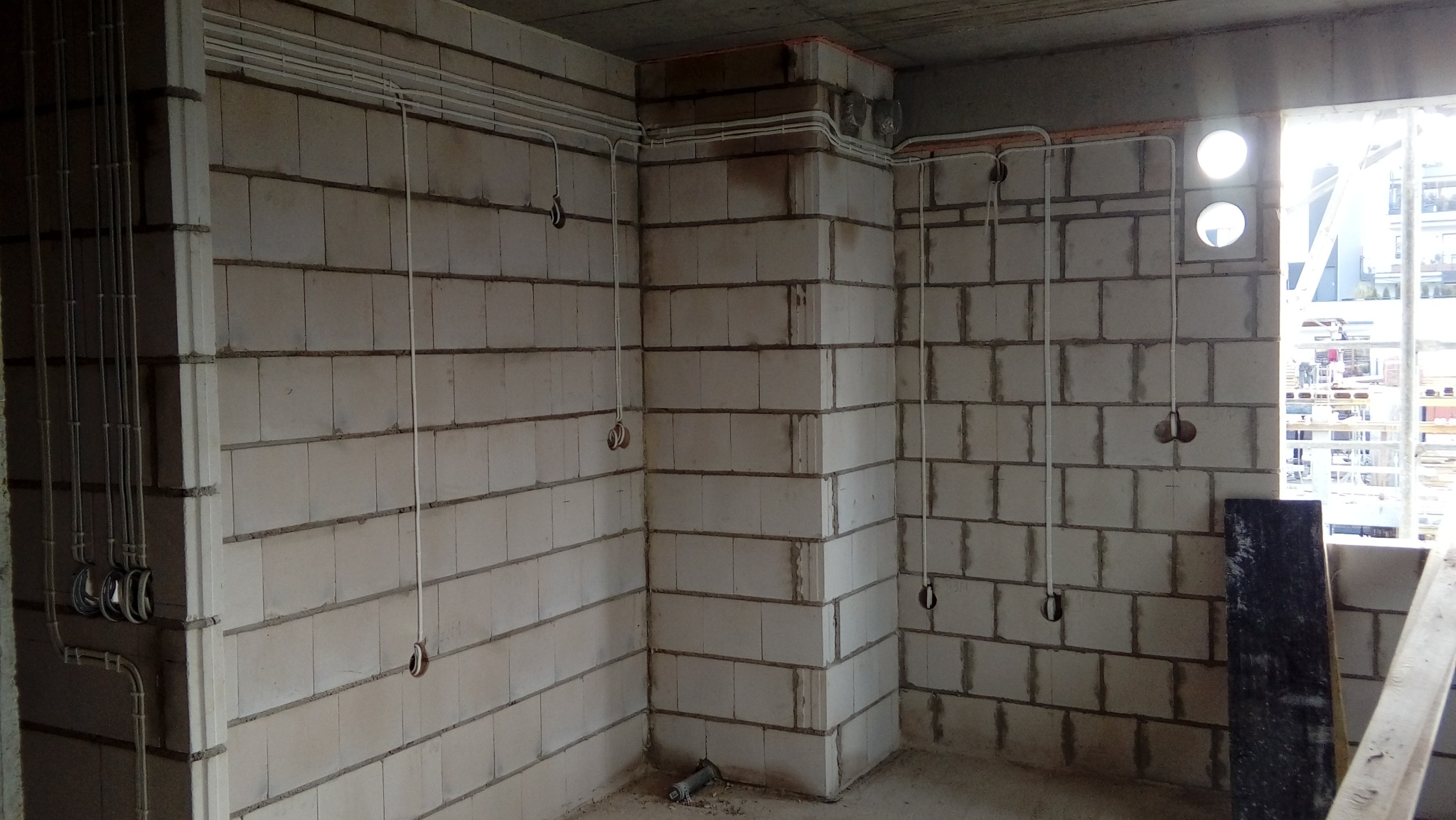 Surowy stan instalacji elektrycznej w pomieszczeniu z białych bloczków, widoczne kable i puszki podtynkowe, w tle okno z widokiem na budowę.