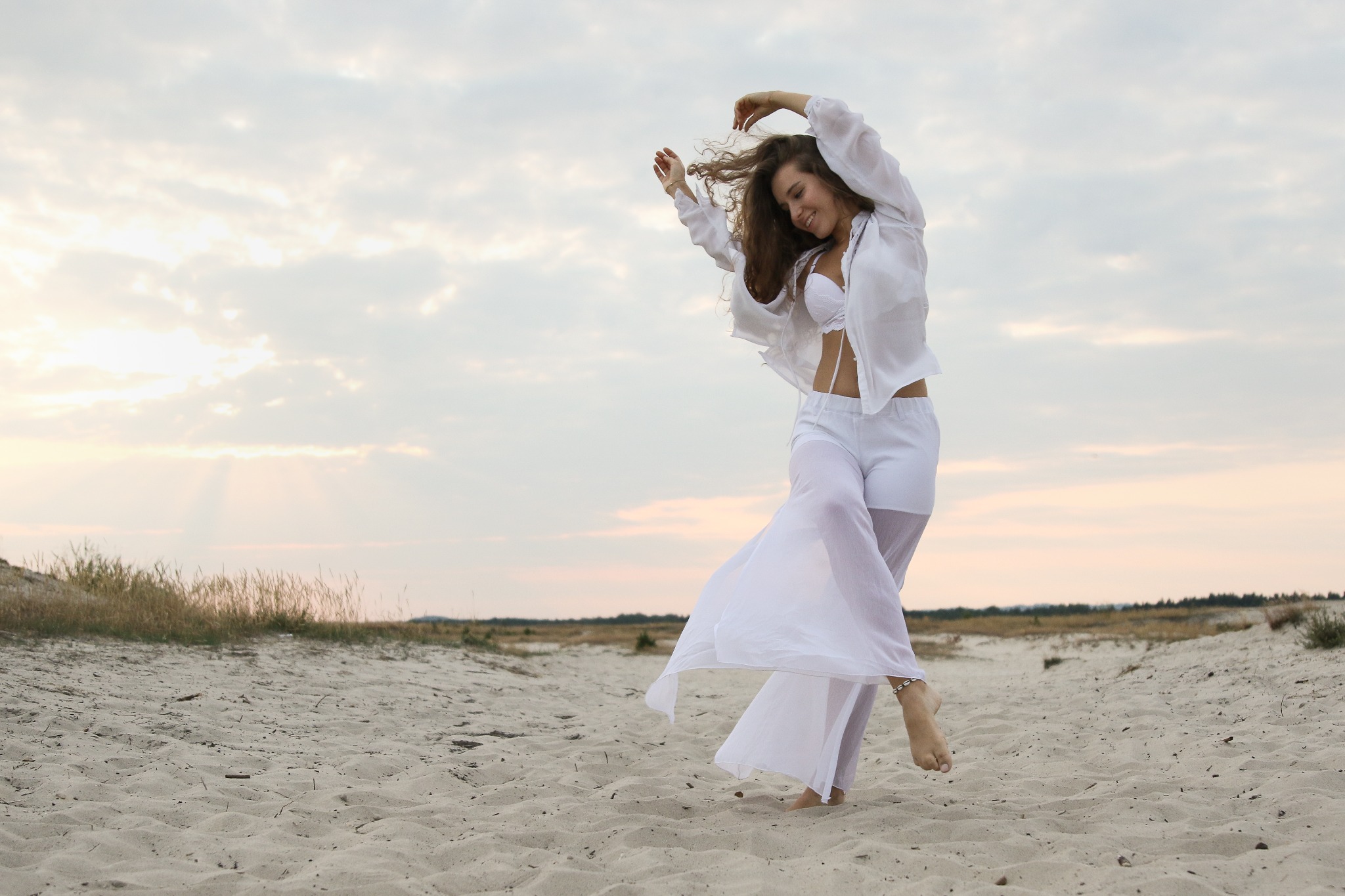 Kobieta w białym stroju tanecznym na piaszczystej plaży, sylwetka w ruchu na tle zachodzącego słońca, delikatne światło podkreśla zwiewność materiału.