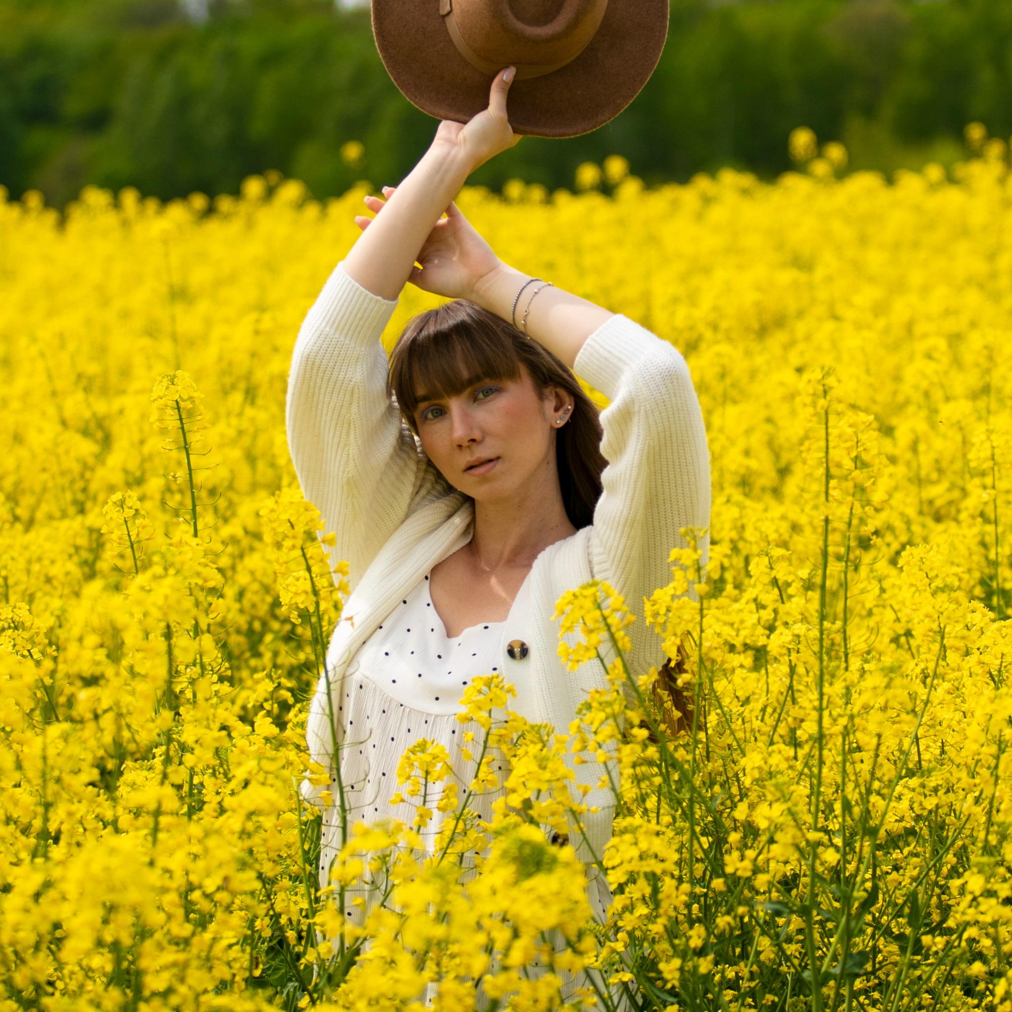 Portret kobiety w kwitnącym polu rzepaku, trzymającej brązowy kapelusz nad głową, ubrana w białą sukienkę w czarne kropki i kremowy kardigan.
