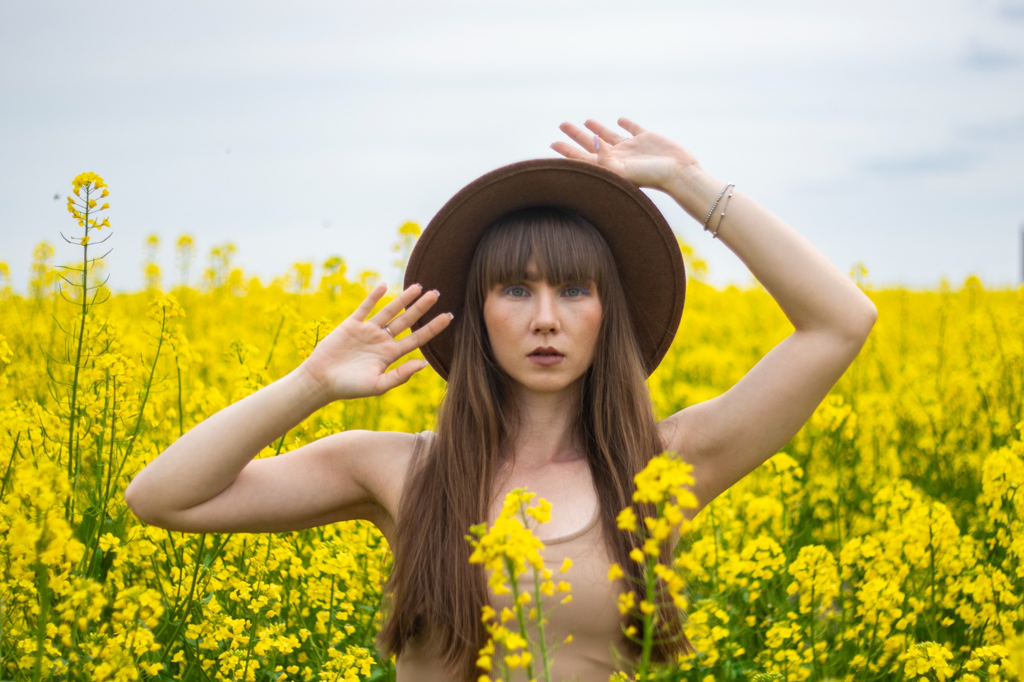 Portret kobiety w brązowym kapeluszu na tle pola kwitnącego rzepaku, z rękami uniesionymi nad głową i delikatnym uśmiechem.