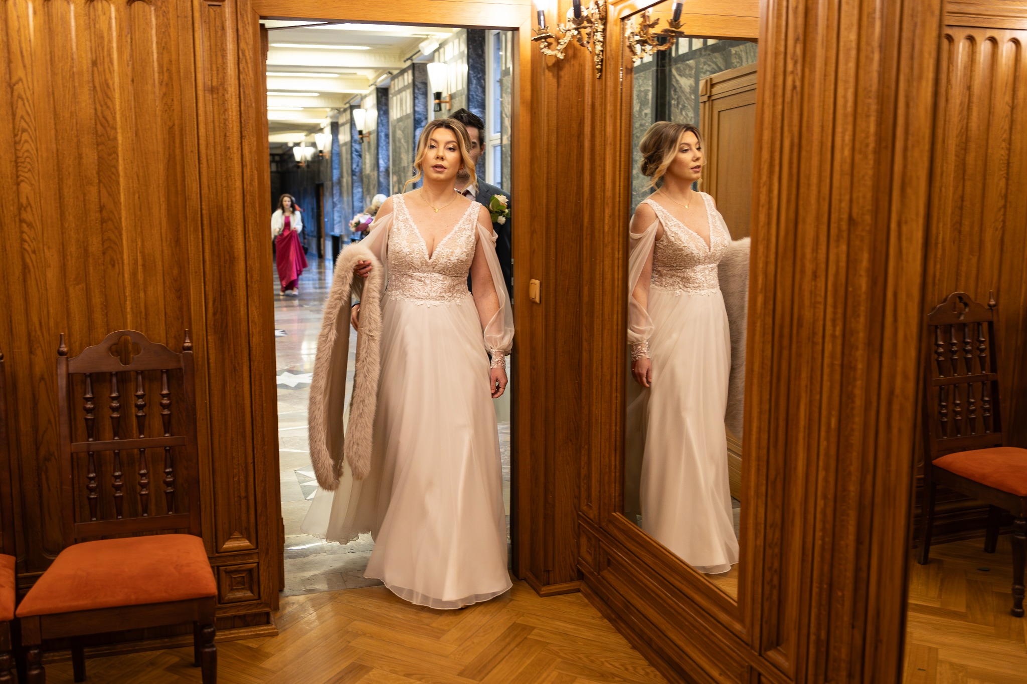 Panna młoda w białej sukni z koronkową górą i futrzanym szalem, wchodząca do sali przez drewniane drzwi, widoczna w lustrze, w tle goście.