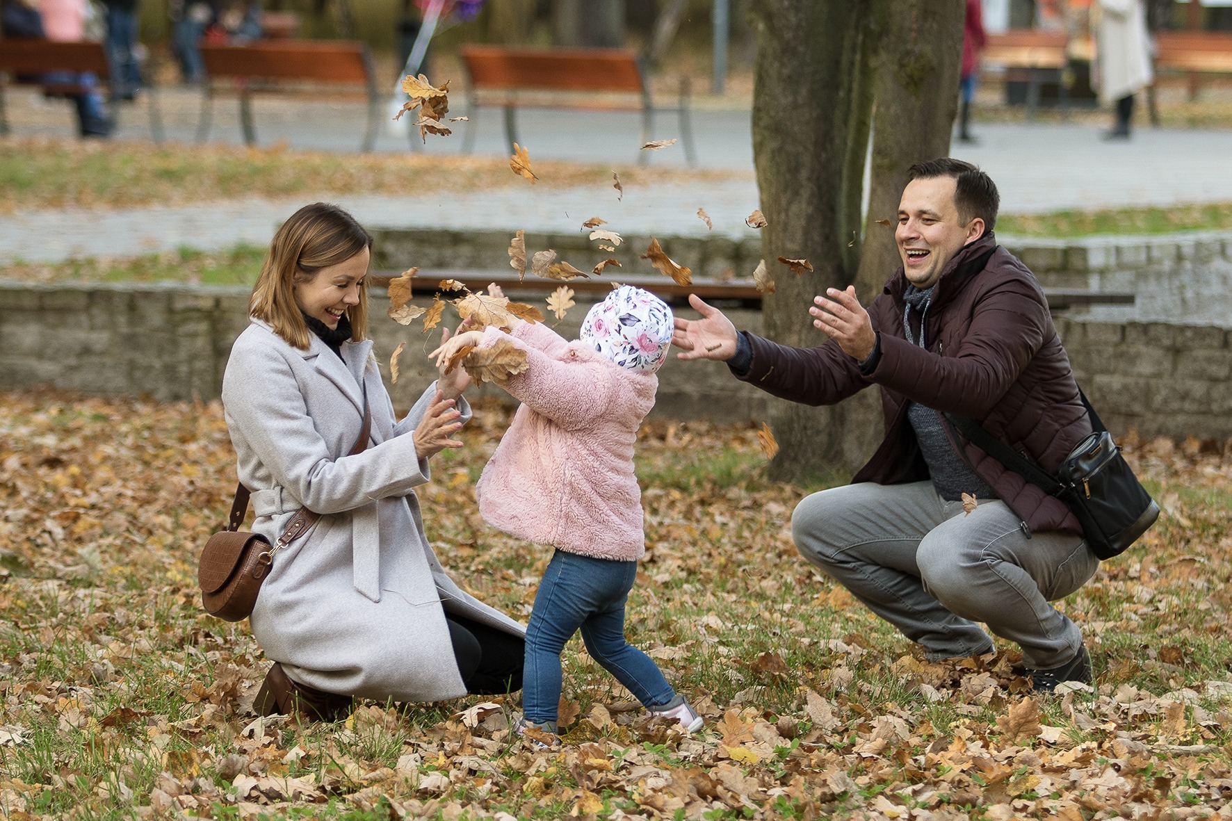 Rodzina bawi się w parku rzucając liśćmi: uśmiechnięta mama w szarym płaszczu, tata w brązowej kurtce i mała dziewczynka w różowej kurtce i kwiecistej czapce. W tle drzewa i ławki parkowe.