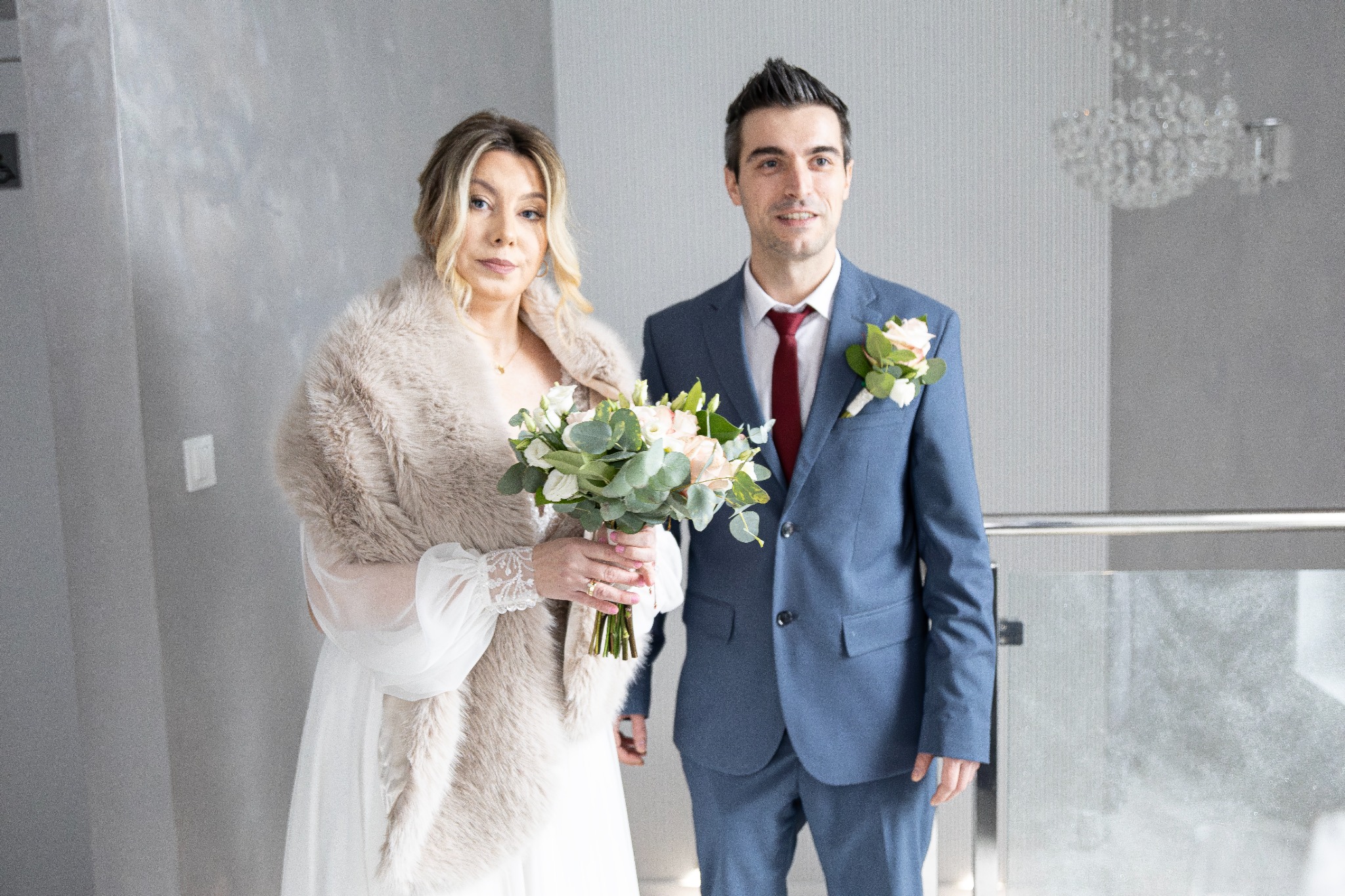 Para młoda w eleganckich strojach ślubnych: panna młoda w futrzanym etolu i bukietem, pan młody w niebieskim garniturze z bordowym krawatem, na tle szarej ściany i balustrady