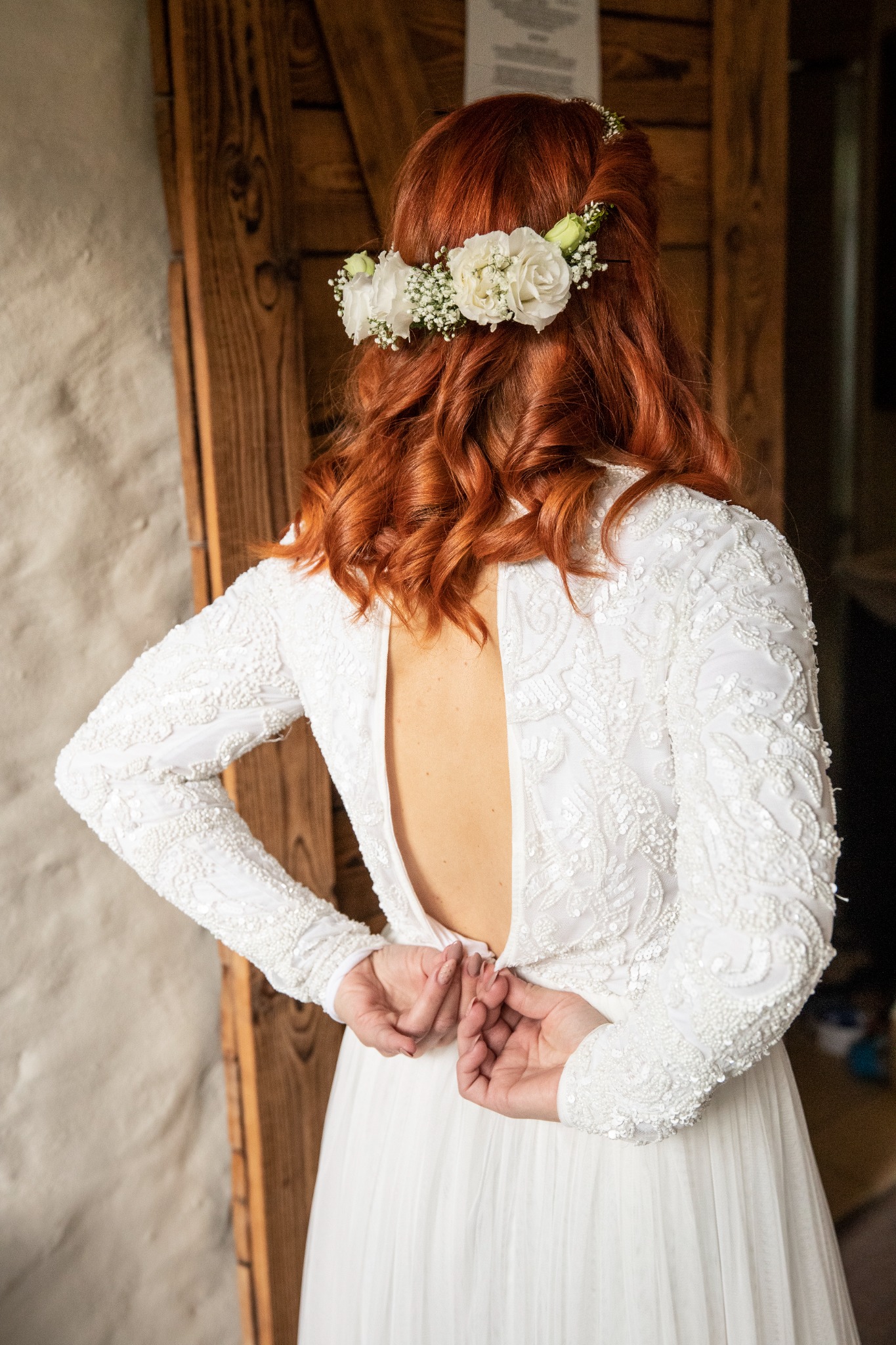 Panna młoda z rudymi włosami i wiankiem z białych kwiatów zapina suknię ślubną z długim rękawem, widok z tyłu na neutralnym tle ściany i drewnianej ramy.