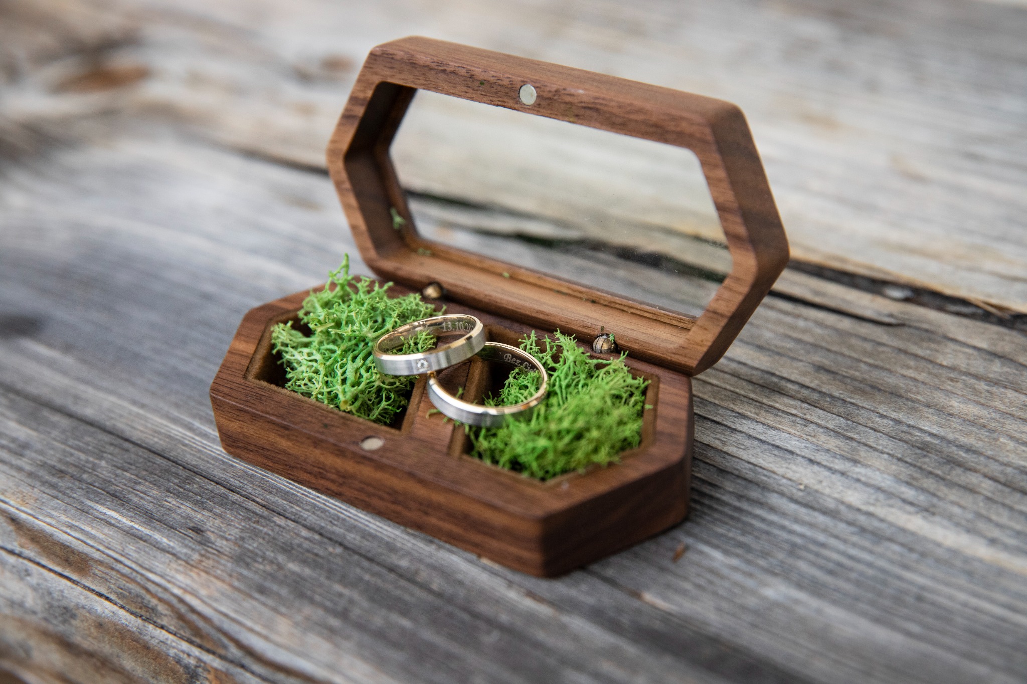 Dwa srebrne obrączki z wygrawerowanymi datami i imionami, leżące w drewnianym, heksagonalnym pudełku z przezroczystym wieczkiem, wypełnionym zielonym mchem, na tle szarego, drewnianego stołu.