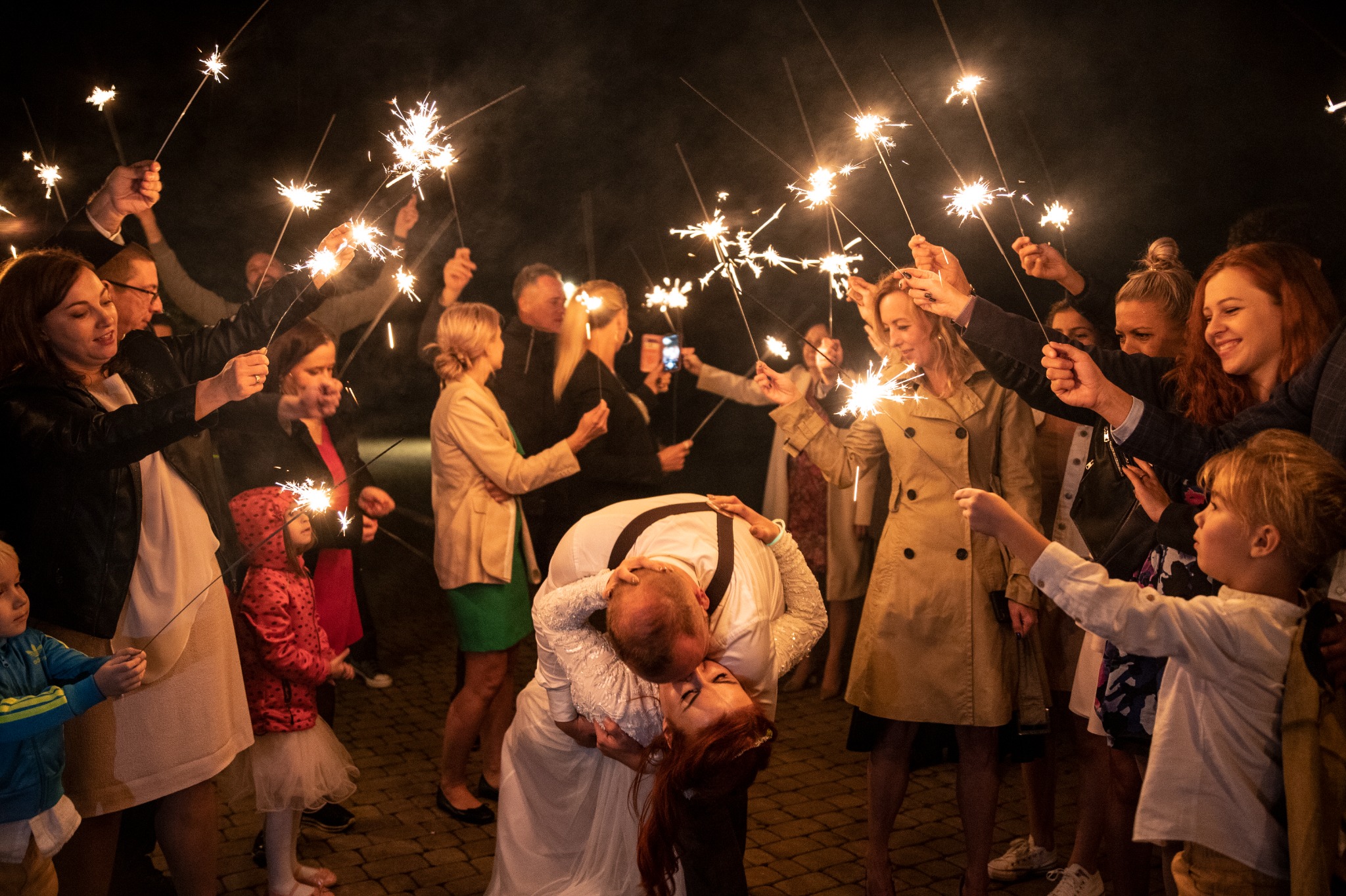 Pocałunek nowożeńców w otoczeniu gości weselnych trzymających zapalone zimne ognie, nocne ujęcie.