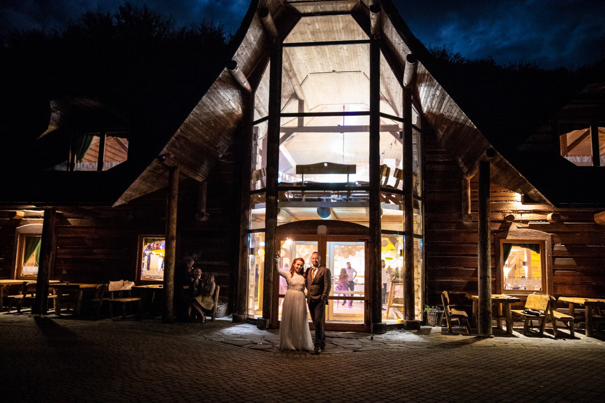 Para młoda stoi przed wejściem do drewnianej sali weselnej w nocy, trzymając zimne ognie. W tle widoczni goście weselni.