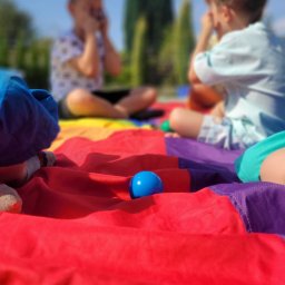 EvenTime - Dzieci bawiące się na kolorowej chuście animacyjnej z niebieską piłką na pierwszym planie, gra na świeżym powietrzu w słoneczny dzień.