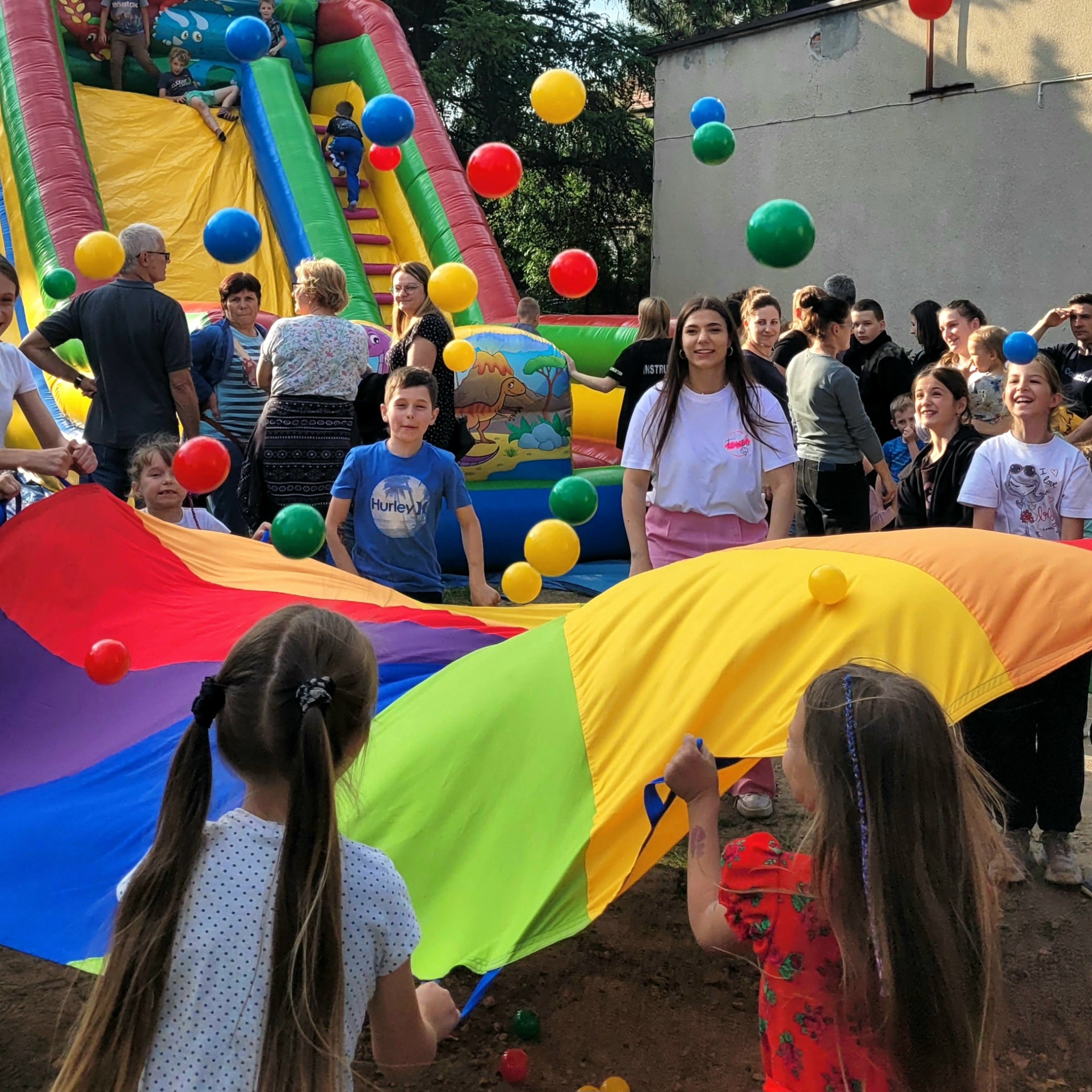 Grupa dzieci i dorosłych bawi się kolorową chustą animacyjną, unosząc w powietrzu kolorowe piłki, w tle dmuchany zamek ze zjeżdżalnią.