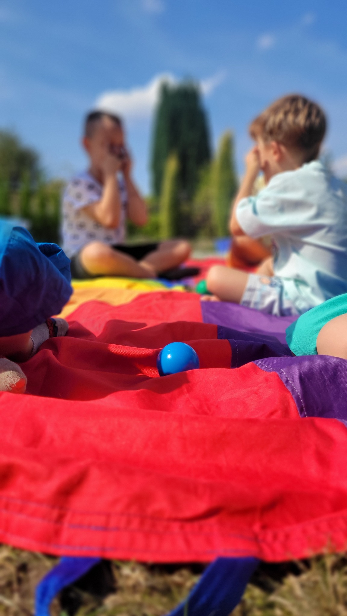Dzieci bawiące się na kolorowej chuście animacyjnej z niebieską piłką na pierwszym planie, gra na świeżym powietrzu w słoneczny dzień.