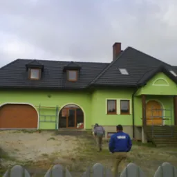 Świeżo otynkowany dom w odcieniach zieleni z ciemną dachówką, widoczne rusztowania i dwaj mężczyźni oddalający się od budynku po zakończonych pracach.