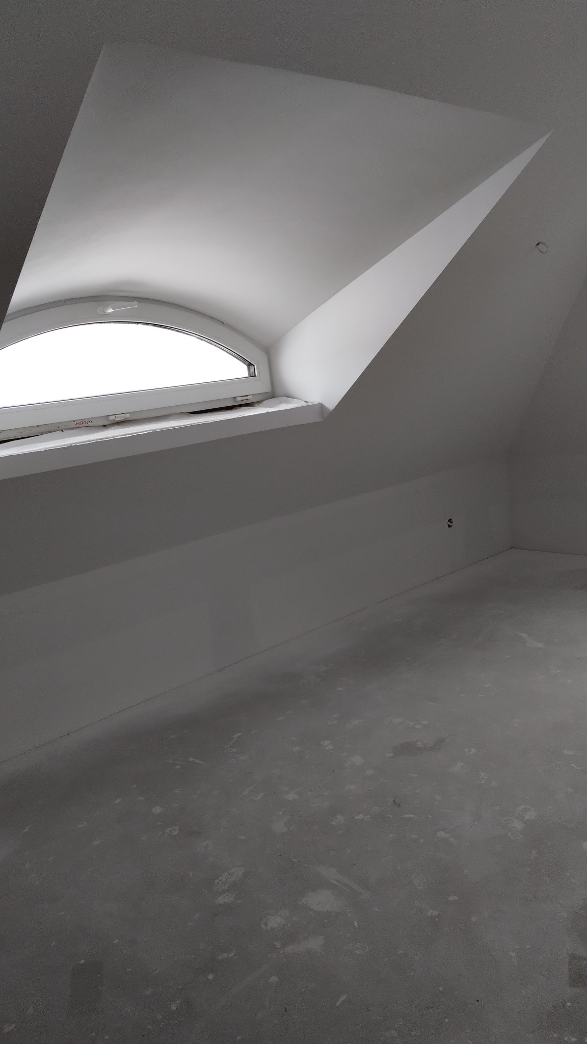 Poddasze z oknem w kształcie łuku, ściany i sufit świeżo pomalowane na biało, surowa podłoga w trakcie przygotowania pod wykończenie.