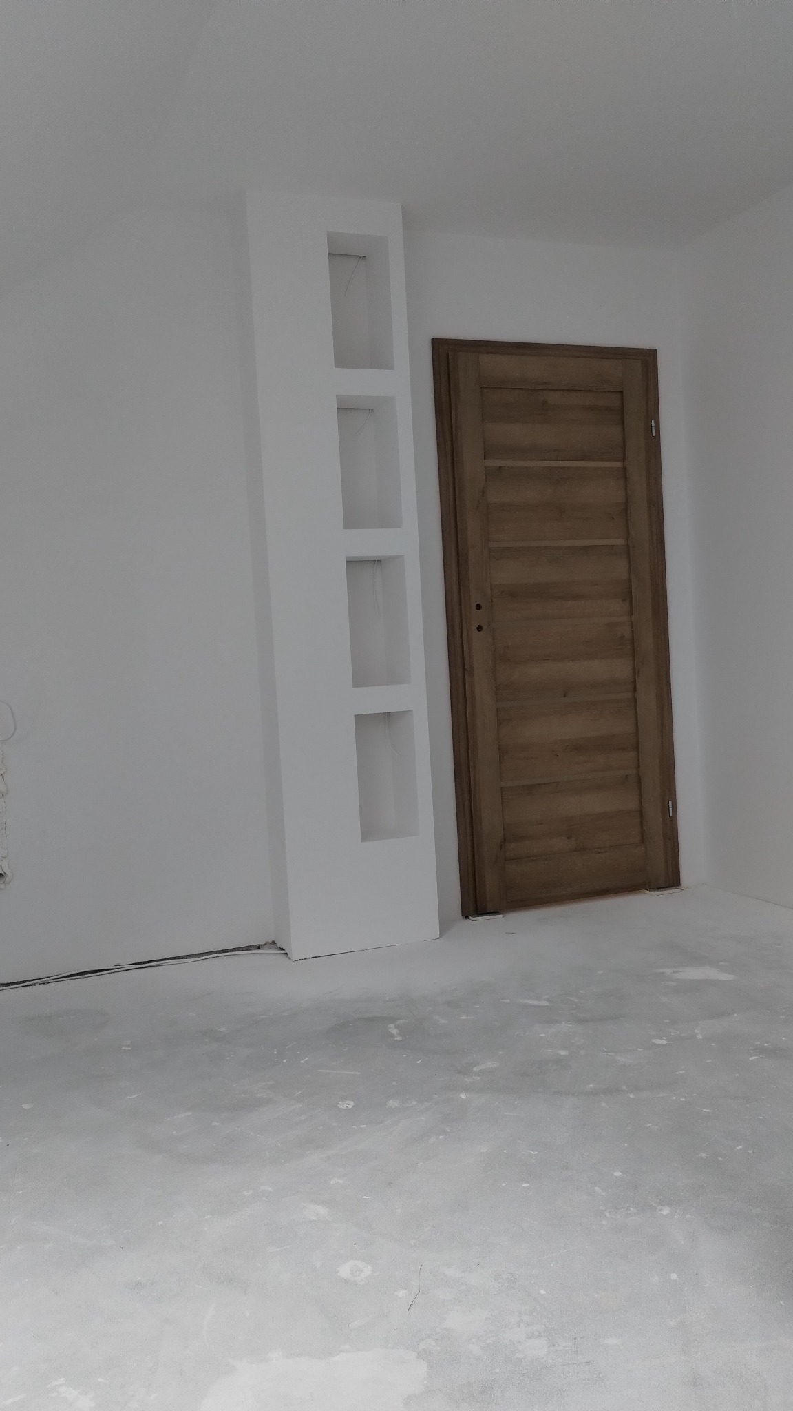 Wnętrze w trakcie prac wykończeniowych: drewniane drzwi wewnętrzne zamontowane obok białej wnęki z półkami, surowa podłoga.
