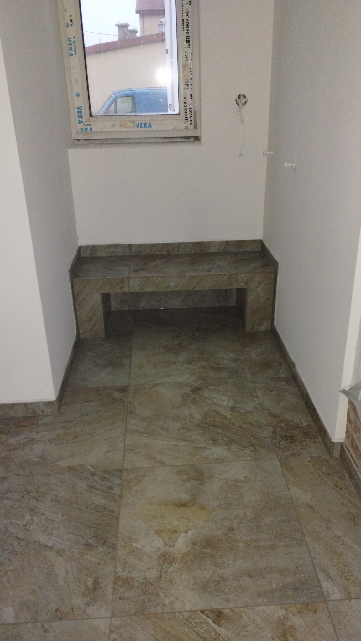 Wykończona wnęka pod oknem z siedziskiem i podłogą wyłożoną płytkami w odcieniach beżu i brązu, widoczny fragment okna z ramą PCV i niebieski samochód za oknem.