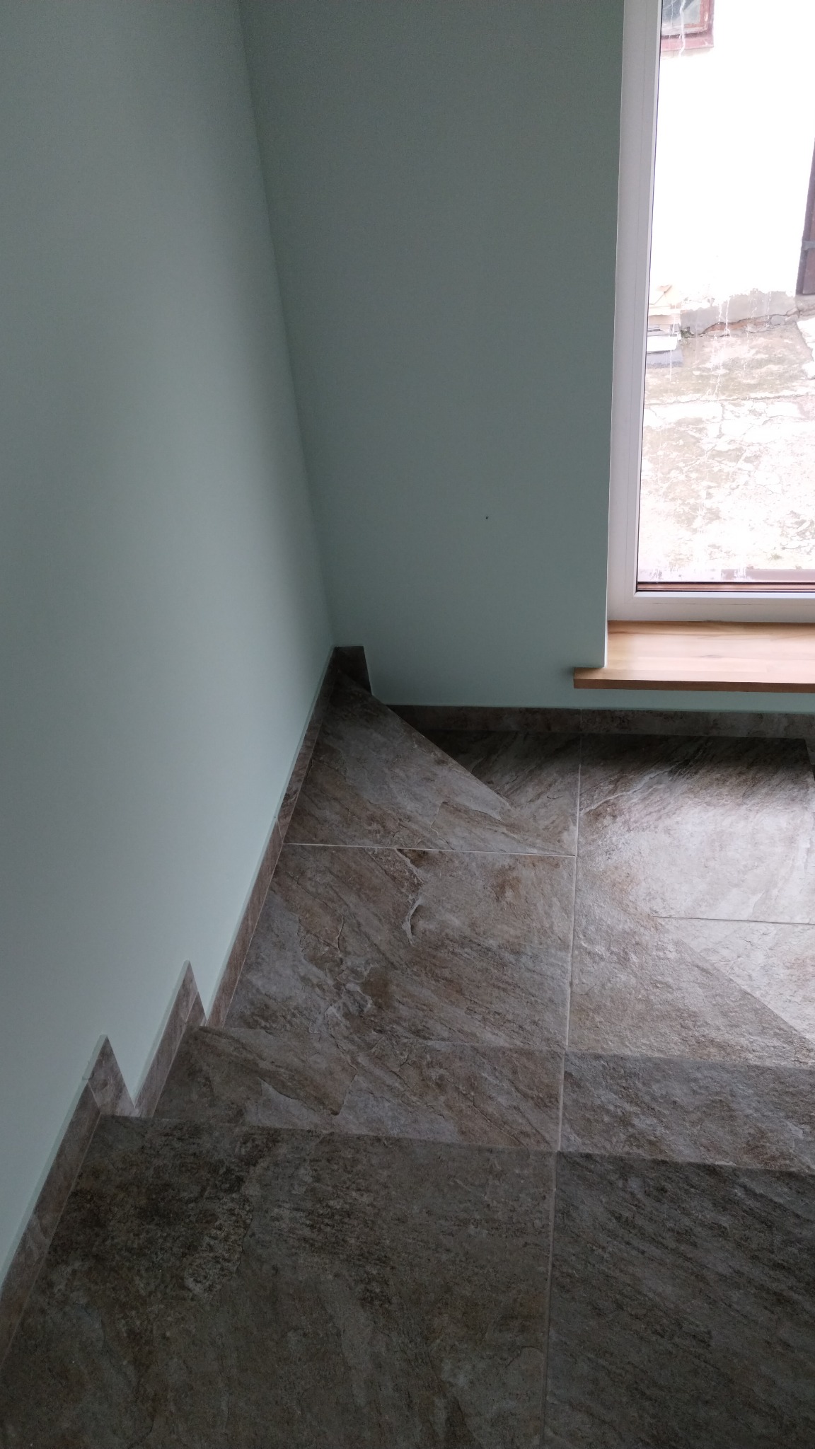 Wykończone schody z brązowych płytek imitujących kamień, z widoczną krawędzią schodów i fragmentem okna z drewnianym parapetem oraz jasną ścianą.