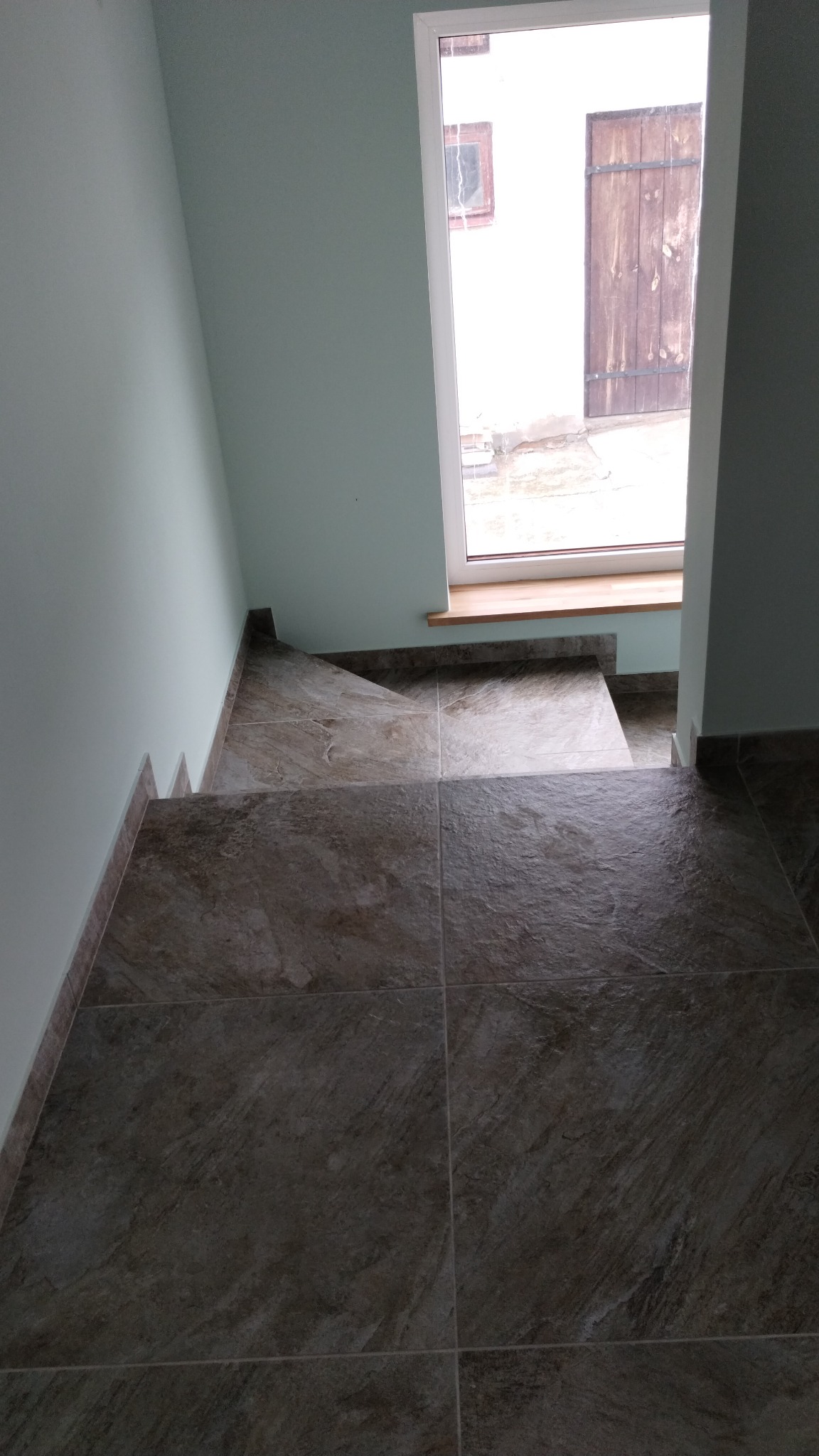 Schody wyłożone ciemnymi, marmurkowymi płytkami ceramicznymi prowadzące w dół, z oknem na półpiętrze z widokiem na budynek gospodarczy z drewnianymi wrotami.