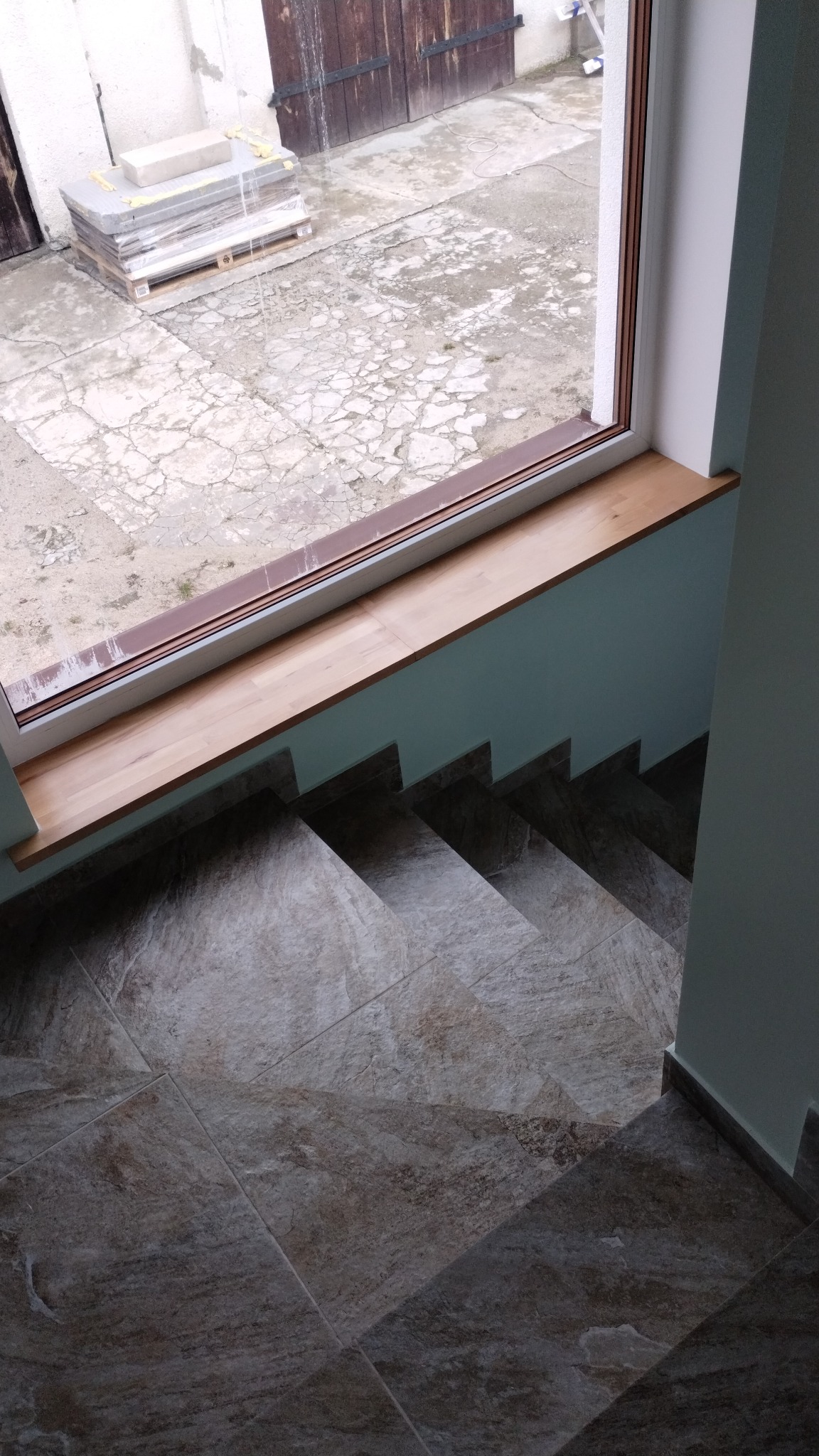 Schody wyłożone płytkami ceramicznymi w odcieniach szarości i brązu, widok z góry, z oknem z drewnianym parapetem w tle, na zewnątrz widoczne płyty budowlane na palecie.