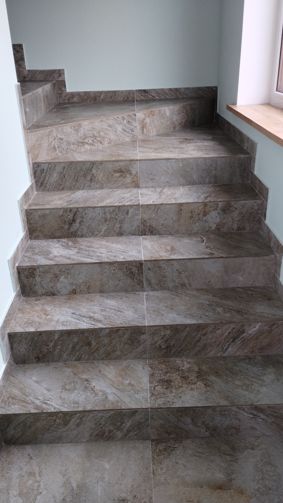 Schody wyłożone brązowo-szarymi płytkami imitującymi kamień, z widocznym oknem i drewnianym parapetem w tle.