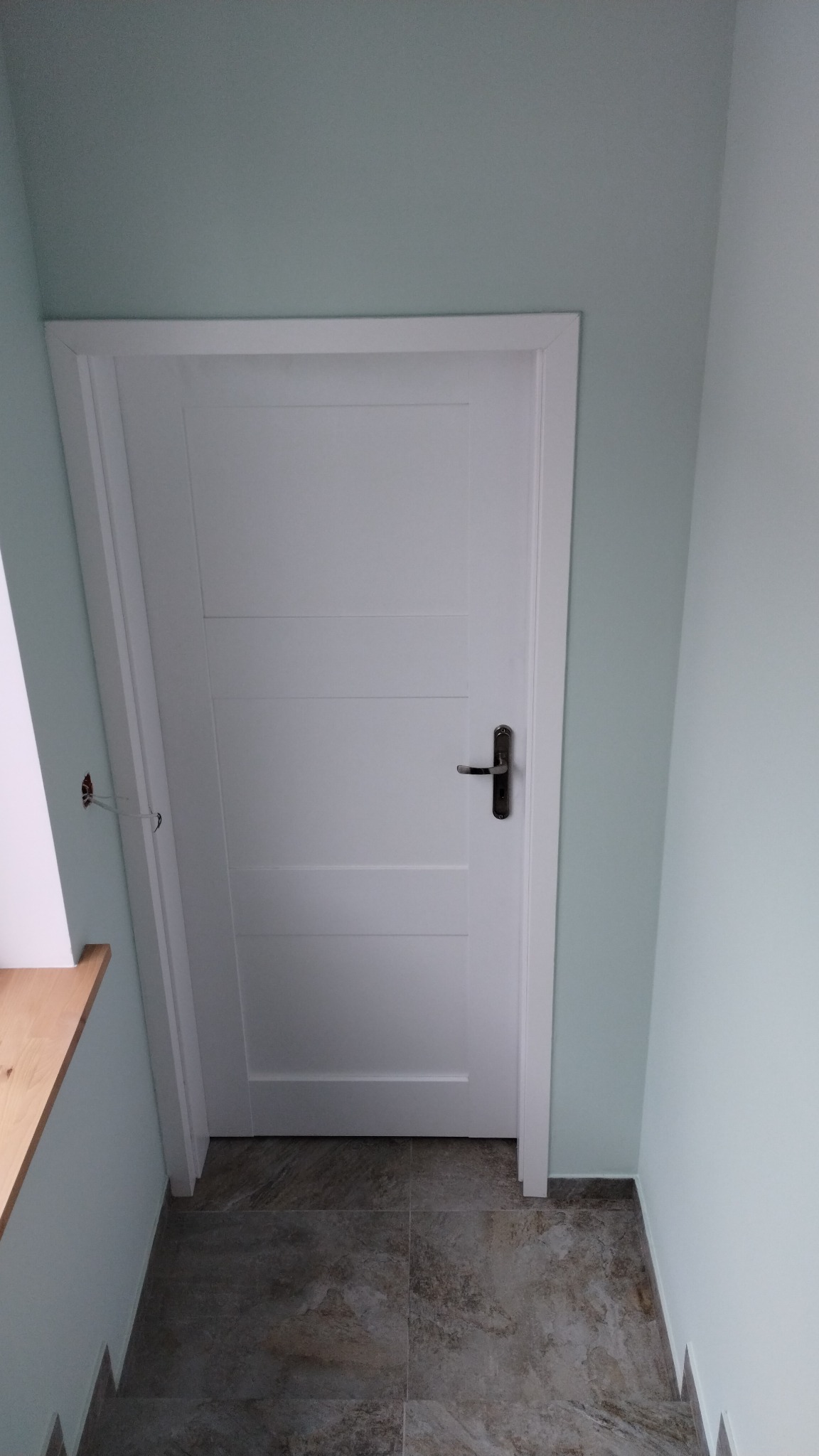 Wnętrze z białą, czteropanelową drzwiami z czarną klamką, szare płytki podłogowe i ściany pomalowane na miętowo. Widoczne wystające kable elektryczne obok futryny.